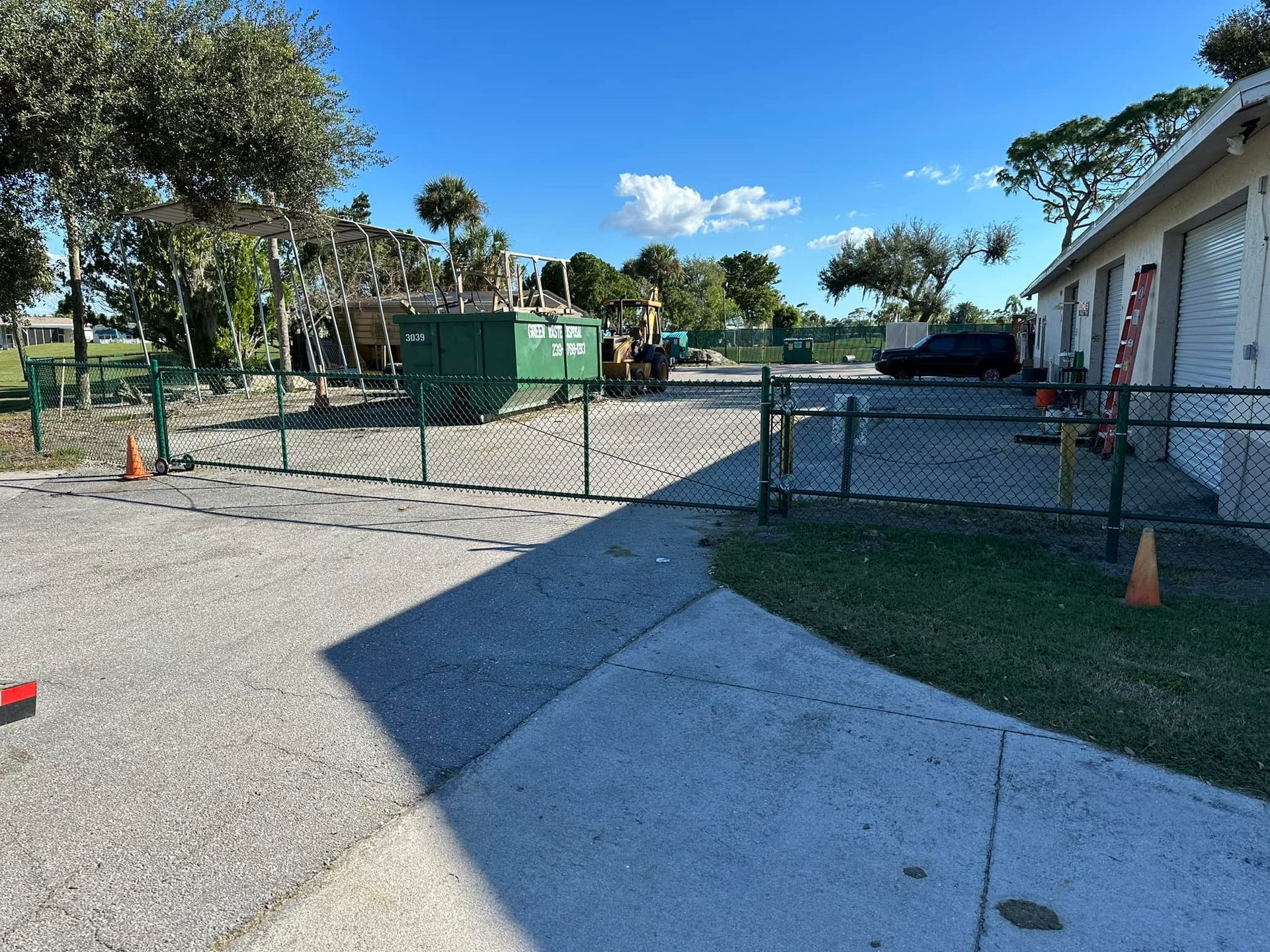 A chain-link fence encloses a gravel area with a dumpster and machinery near a building on a sunny day.