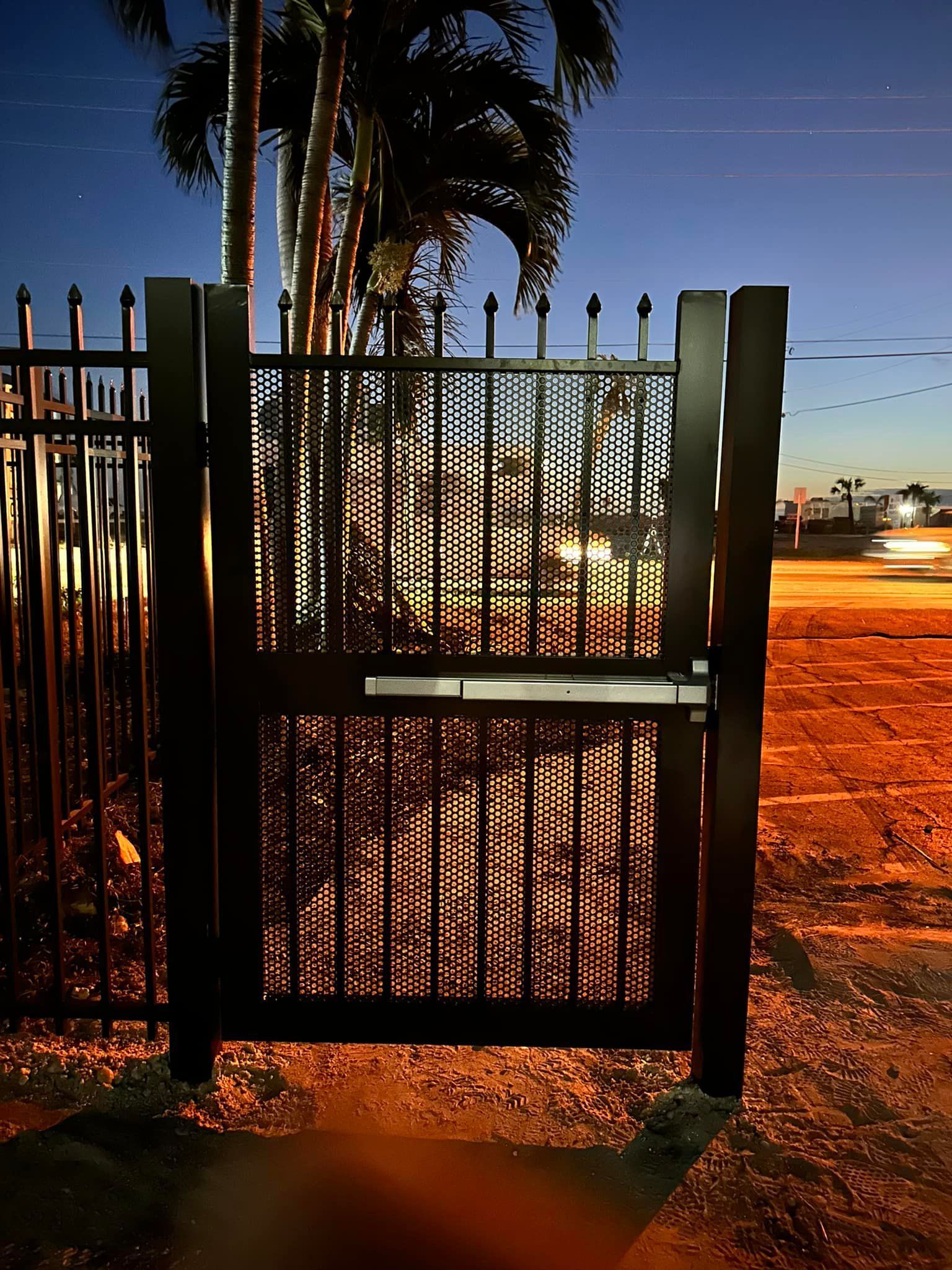 Black metal gate with a decorative pattern at dusk; street and palm trees in background.