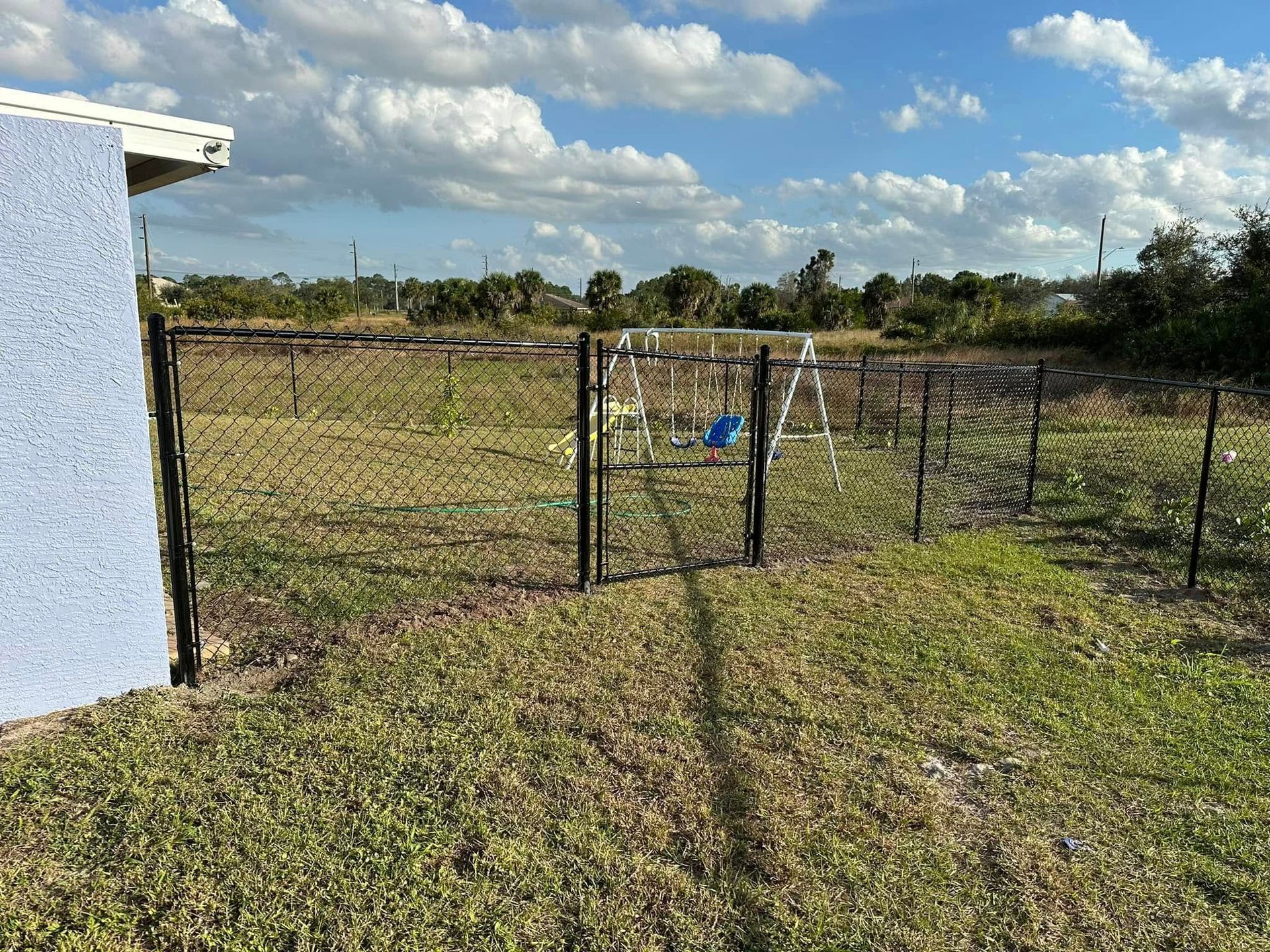 A backyard with a chain-link fence, a swing set, and a blue-painted house under a cloudy sky.