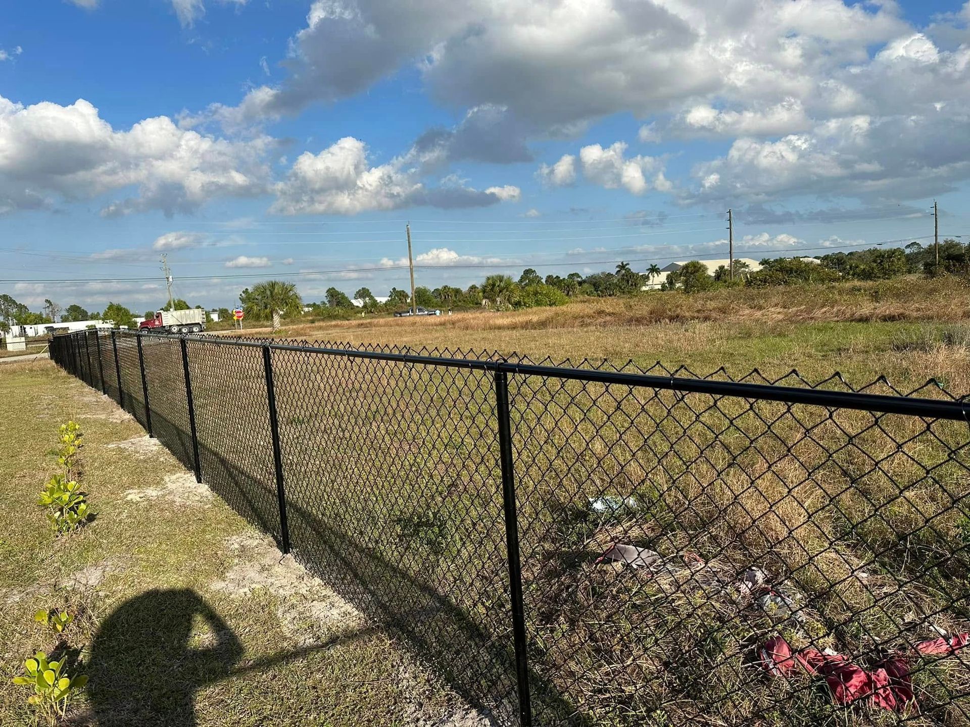 Black chain-link fence bordering a field with scattered debris under a partly cloudy sky.