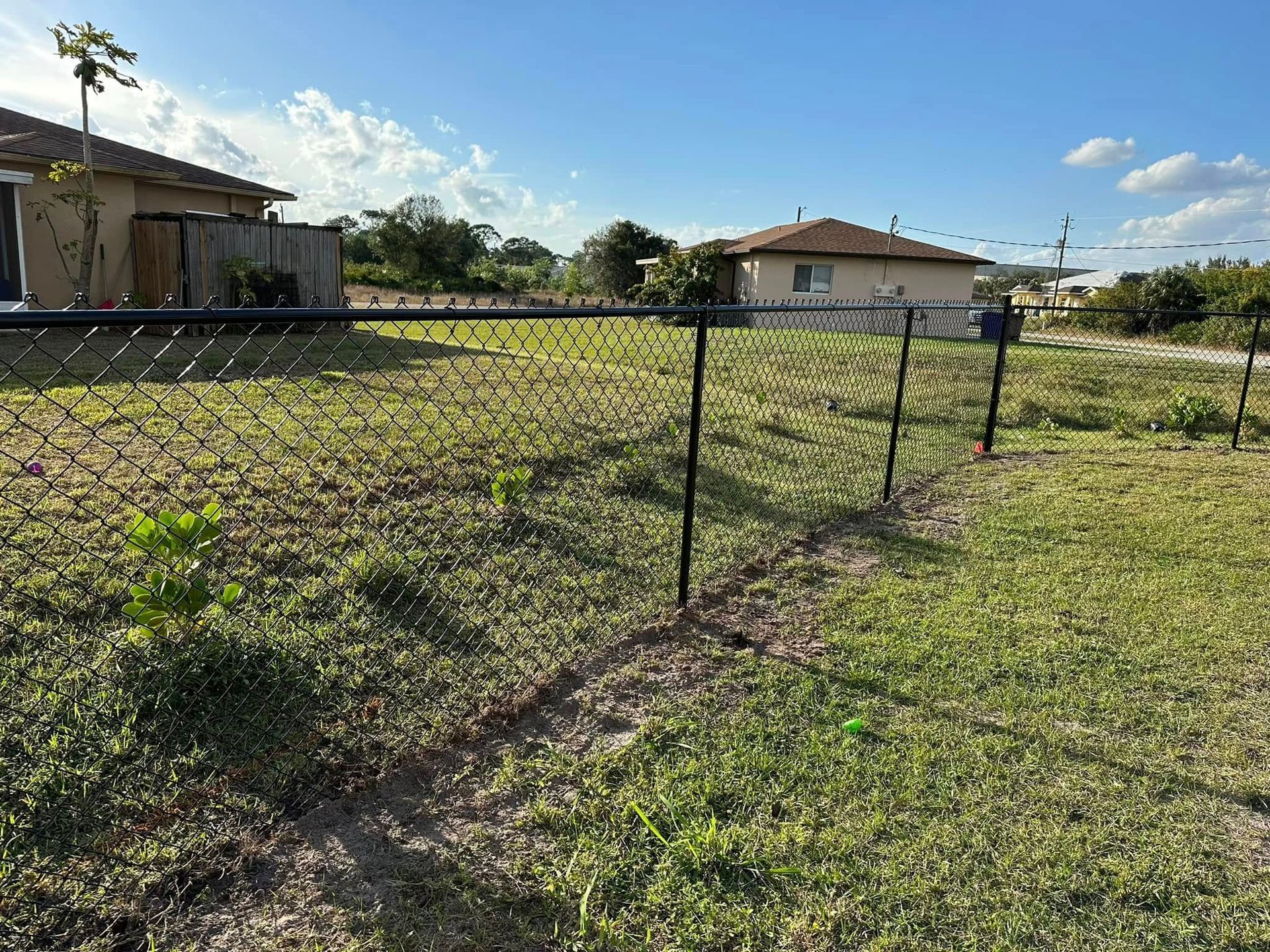Chain-link fence in a grassy backyard with two houses in the background under a blue sky.