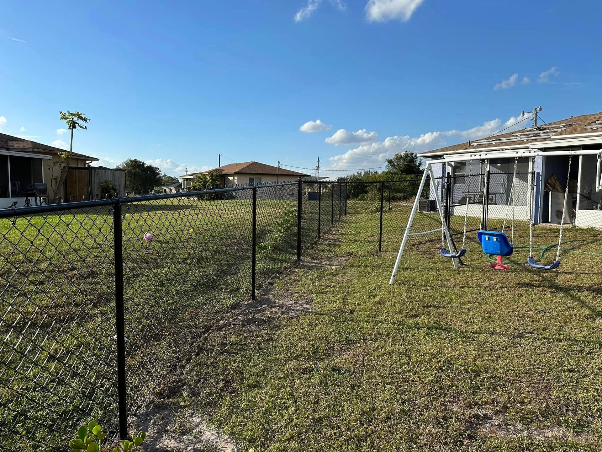 Black chain-link fence in a grassy backyard with a swing set, houses, and a blue sky.