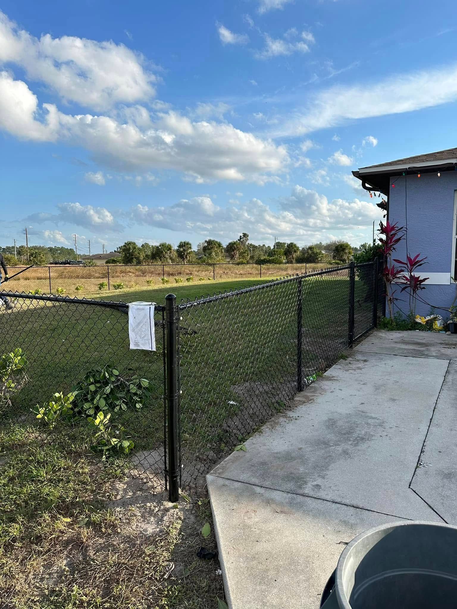 Black chain-link fence next to a concrete walkway. Green field and blue house under a cloudy sky.