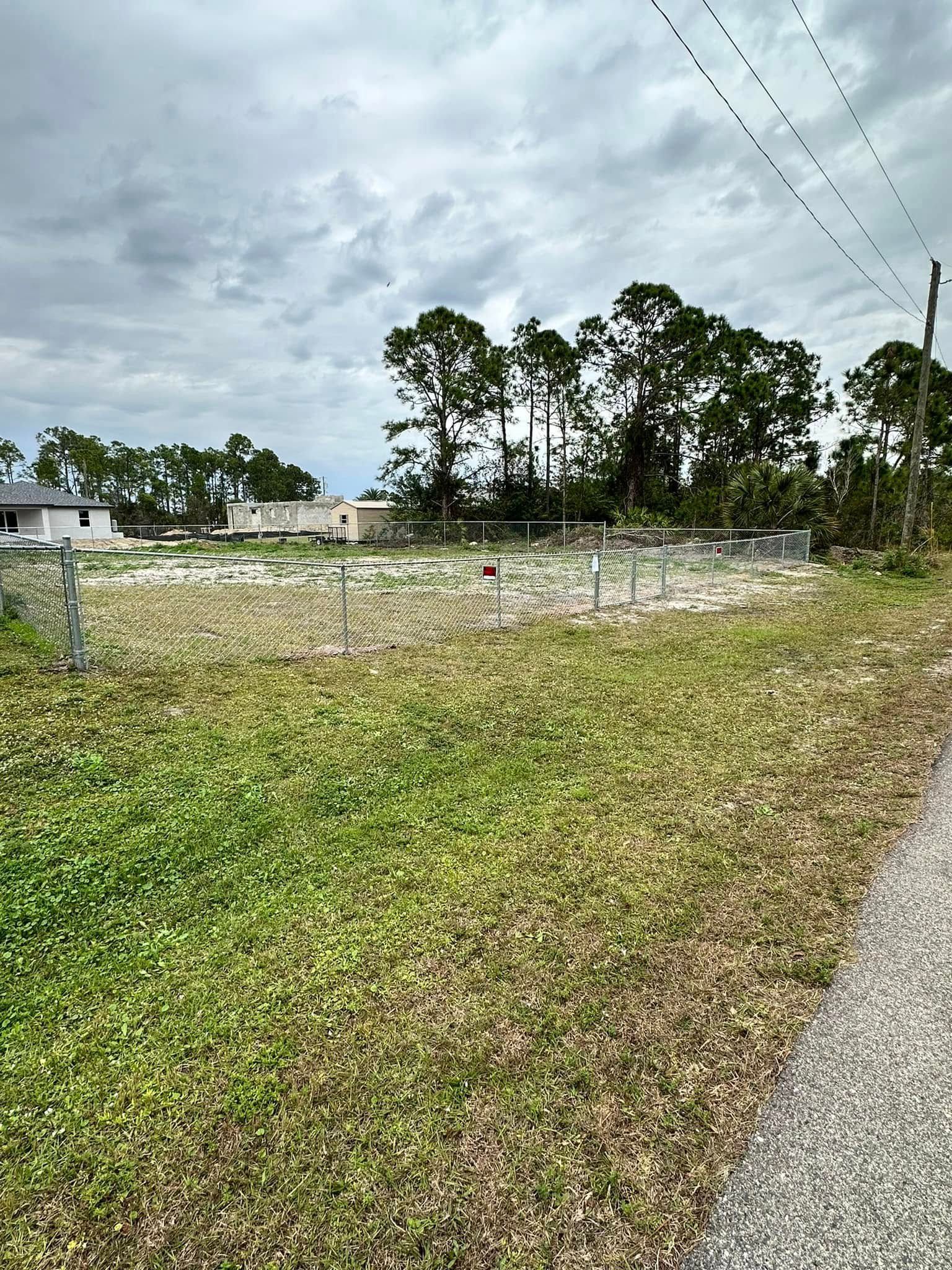 Grassy empty lot with a chain-link fence, trees, and overcast sky. Gravel road in the foreground.