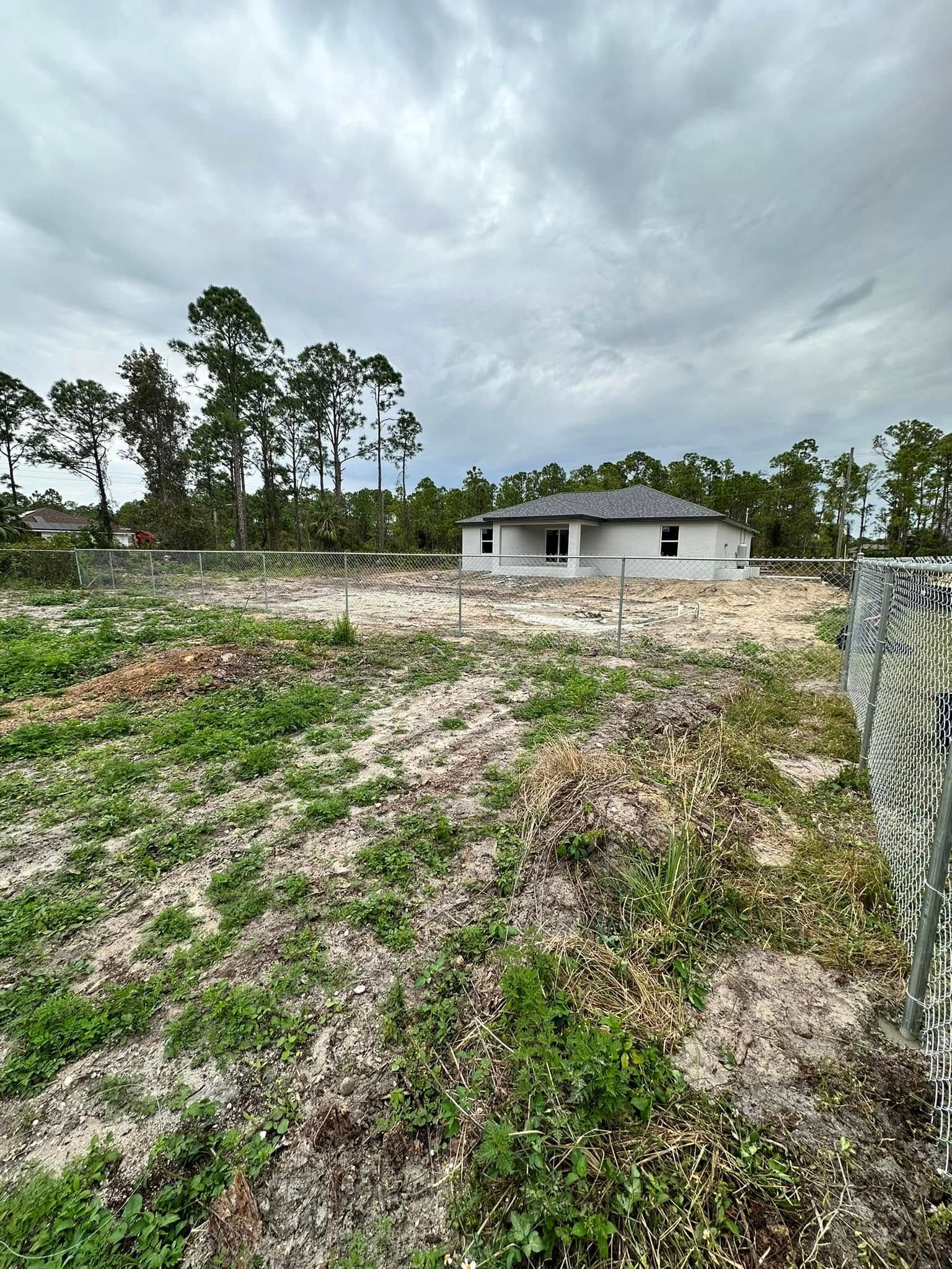 House under construction on a sandy lot with sparse vegetation, surrounded by trees under a cloudy sky.