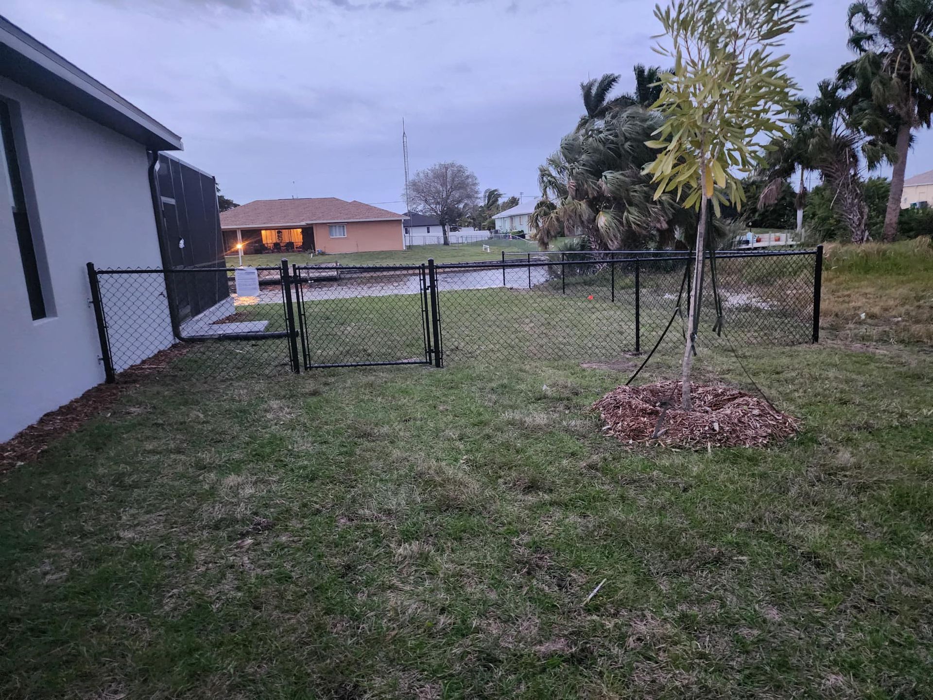 Black chain-link fence encloses a backyard lawn with a young tree, facing a canal.