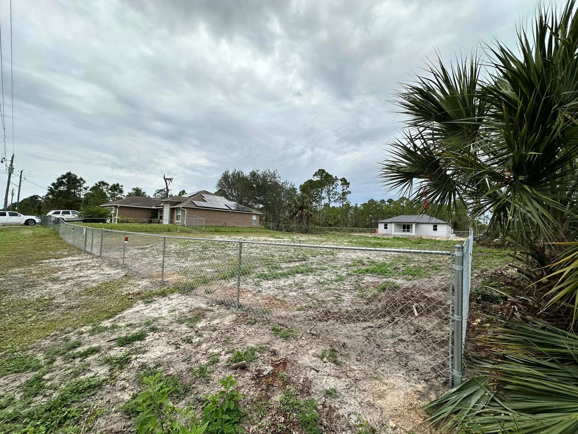 Empty lot with a chain link fence, houses, and cloudy sky.