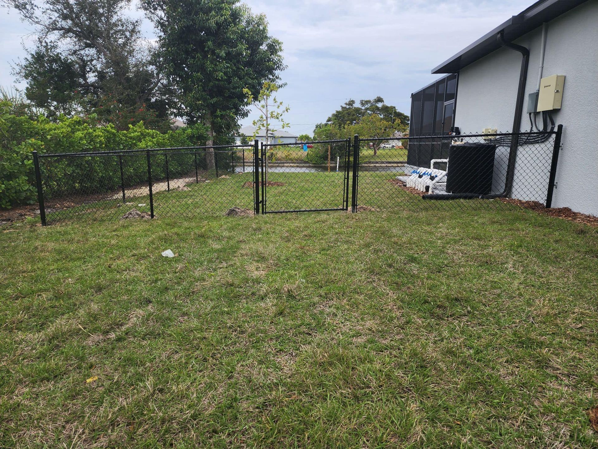Black chain-link fence with a gate in a grassy backyard. House in the background. Overcast day.