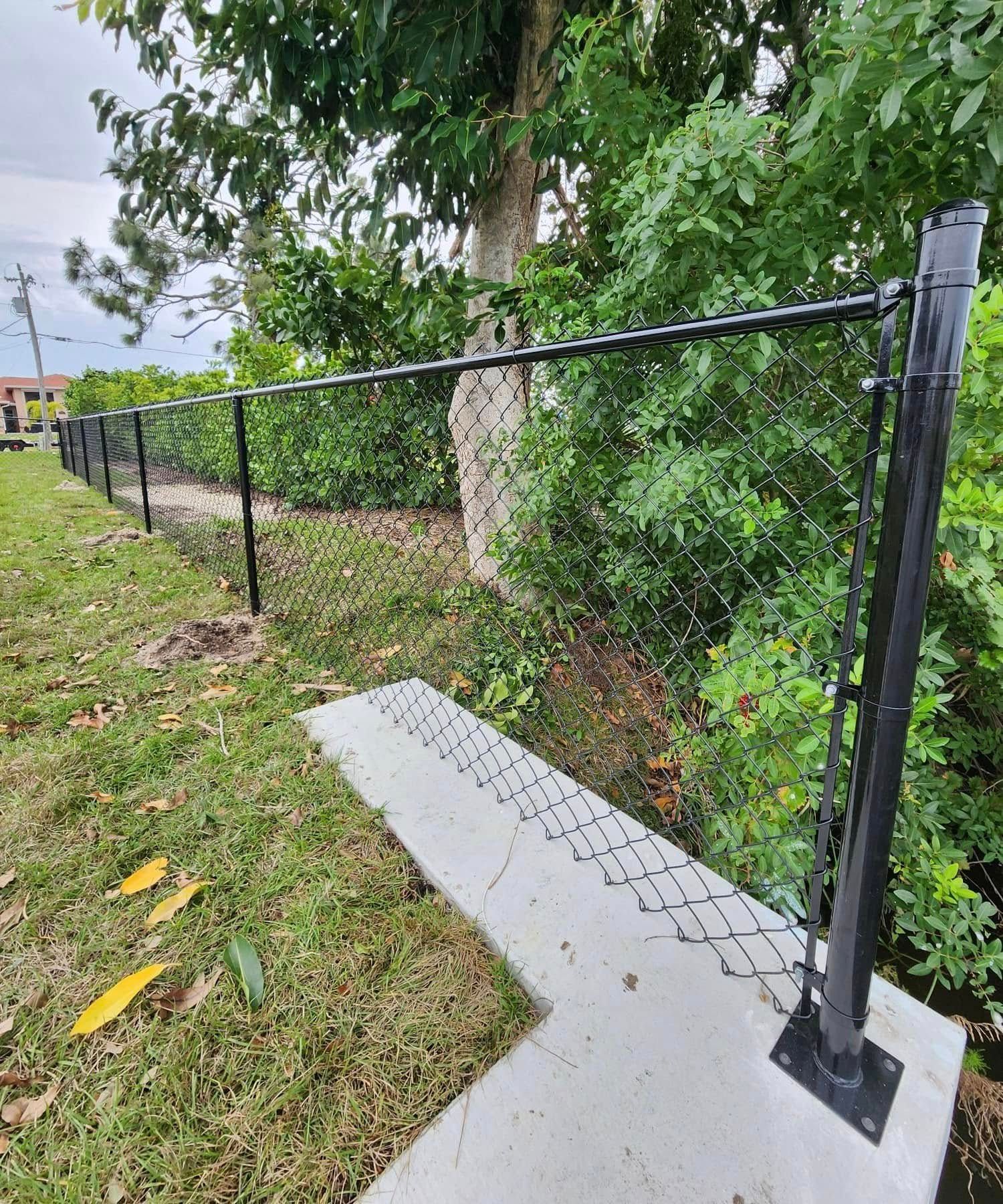 Black chain-link fence alongside a concrete pathway. Green foliage and grass surround it.