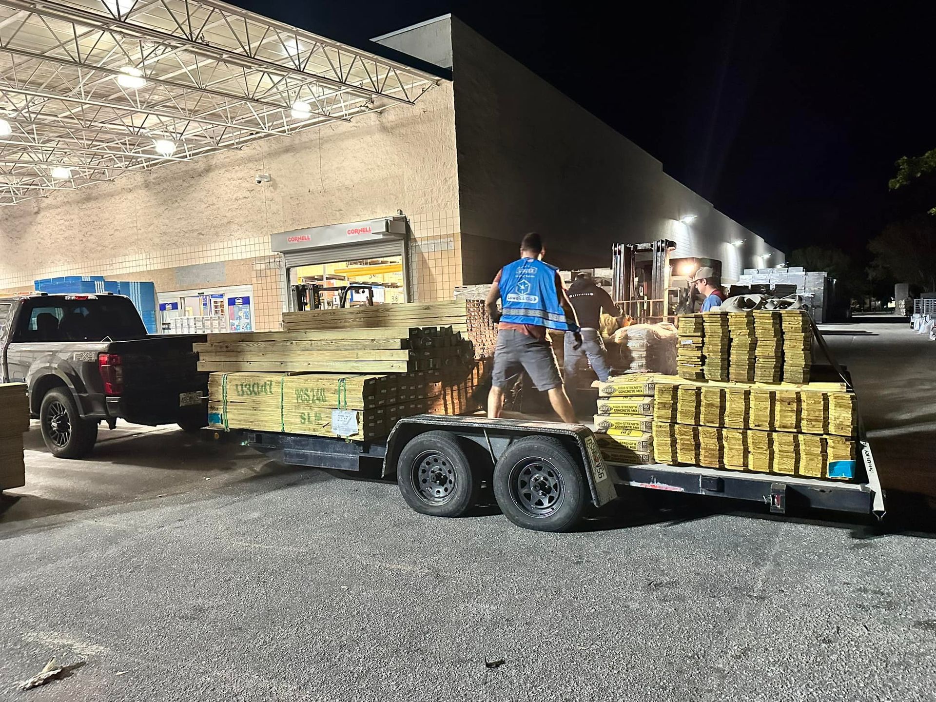 Person loading lumber onto a trailer from a store at night. Truck parked beside.