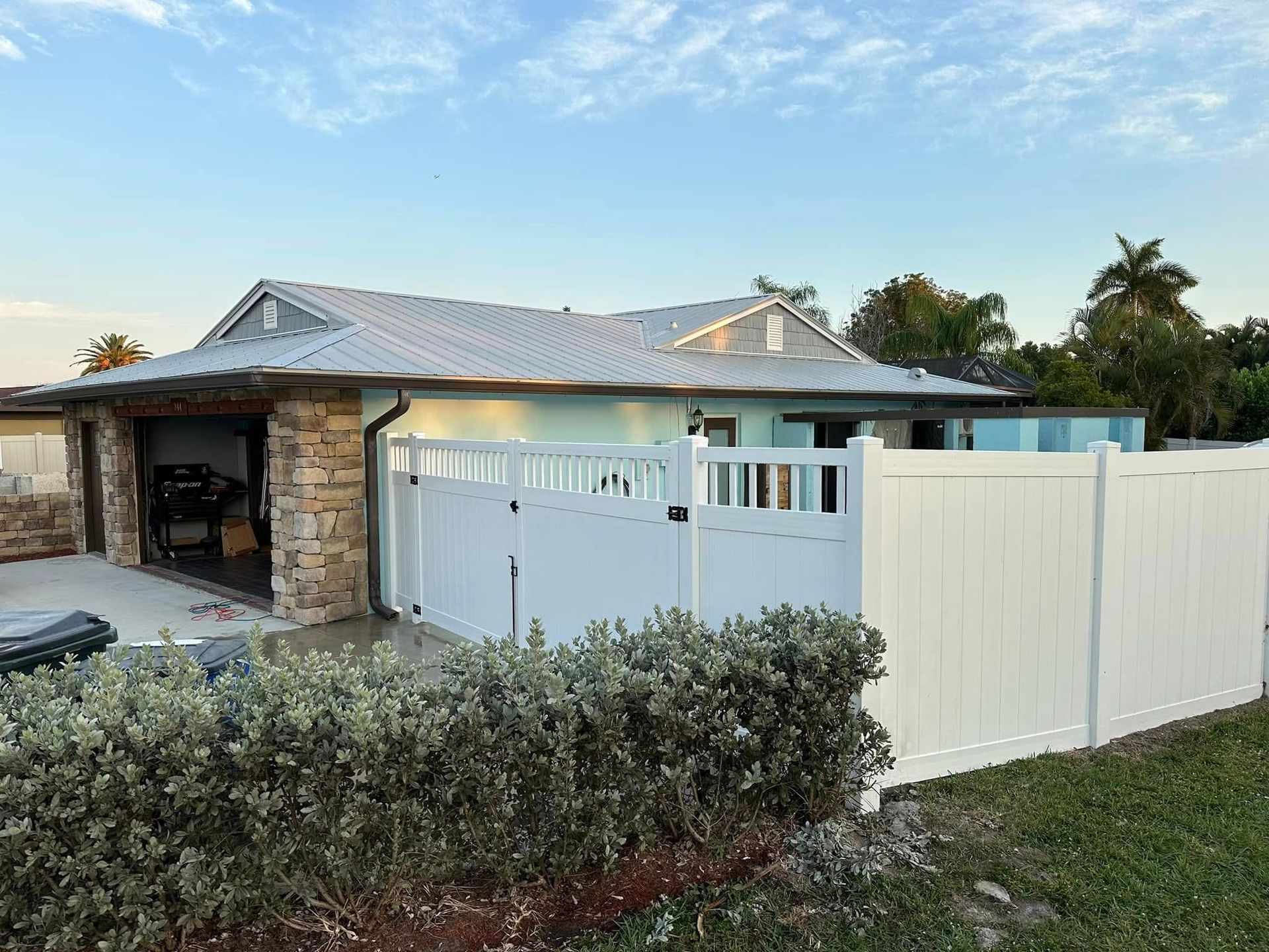 White fence surrounds a house with a stone garage, blue exterior, and green plants under a blue sky.