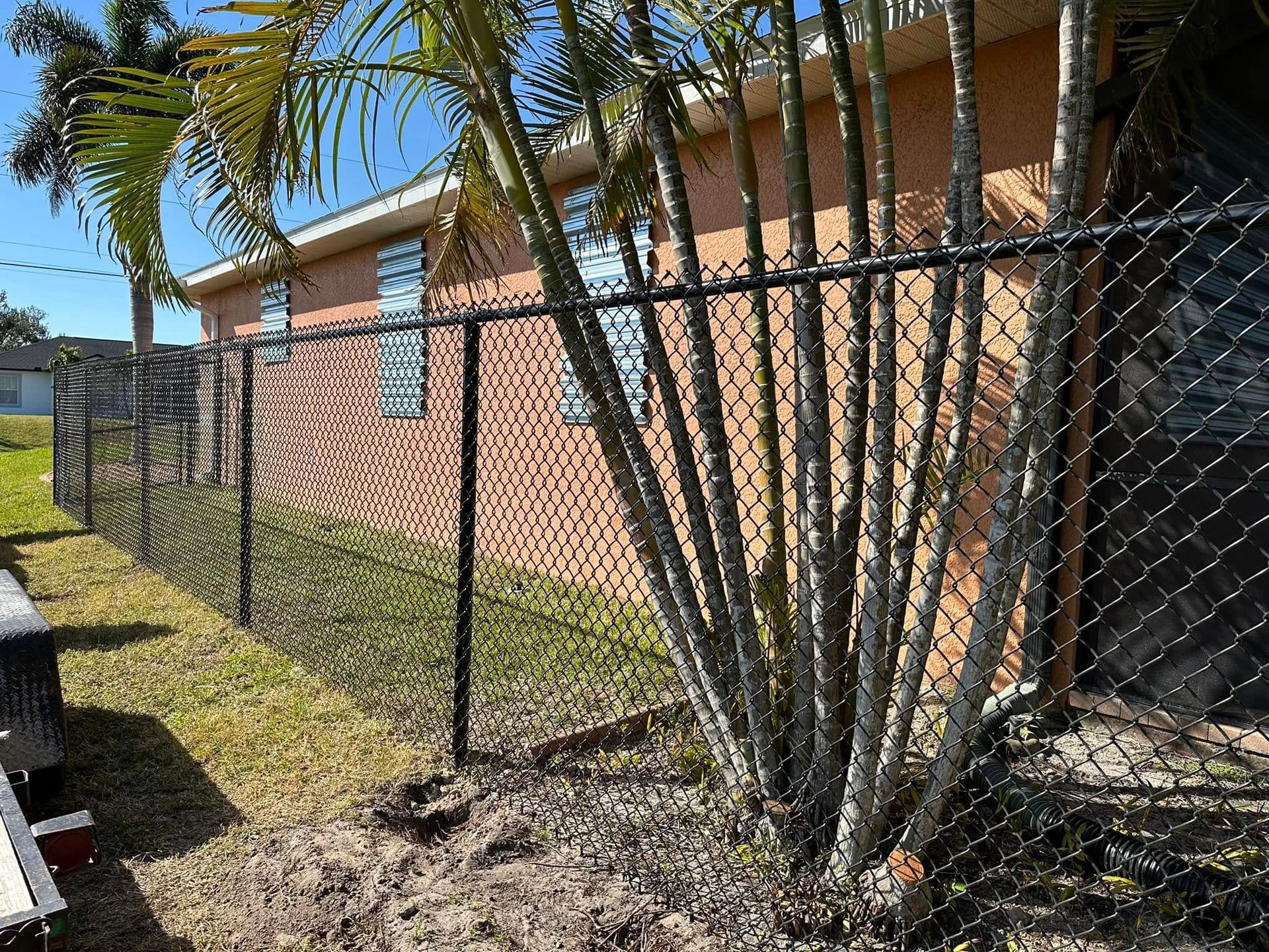 Black chain-link fence runs along a brick building with palm trees nearby on a sunny day.