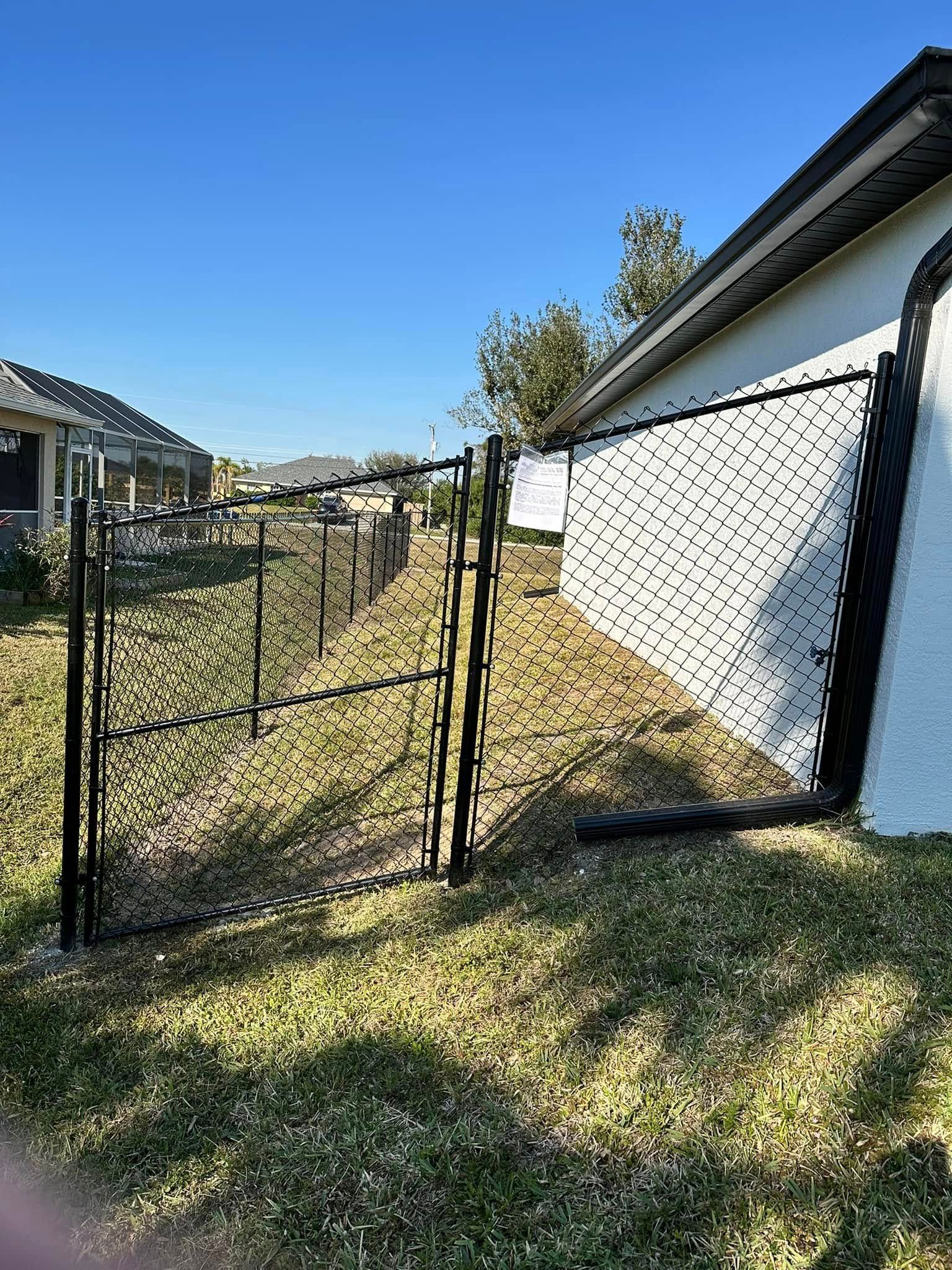 Black chain-link fence with a gate next to a house and grassy area. Blue sky.