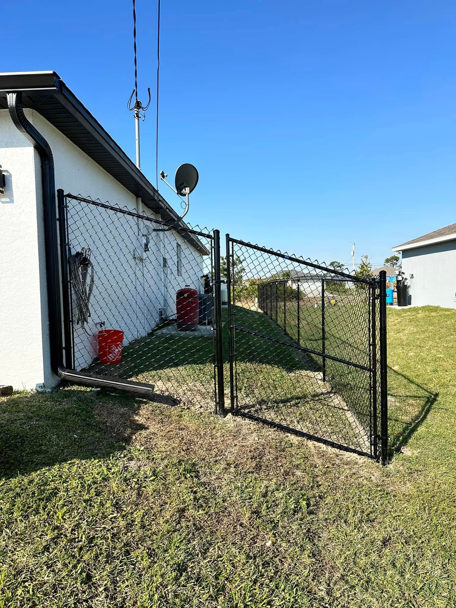 Black fence with gate next to a white house, antenna and satellite dish, red containers, and green grass.