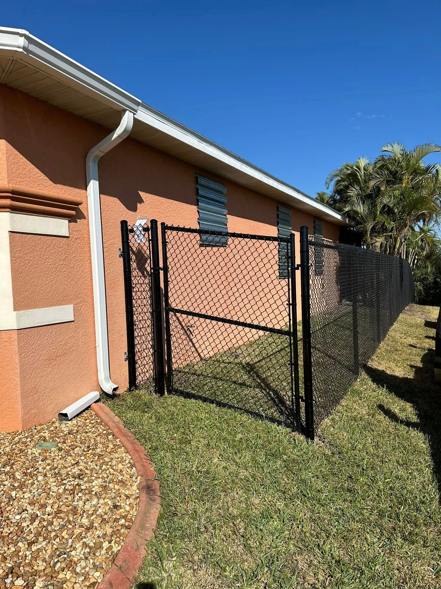 Black gate in a yard next to a stucco building with a white gutter, under a clear blue sky.