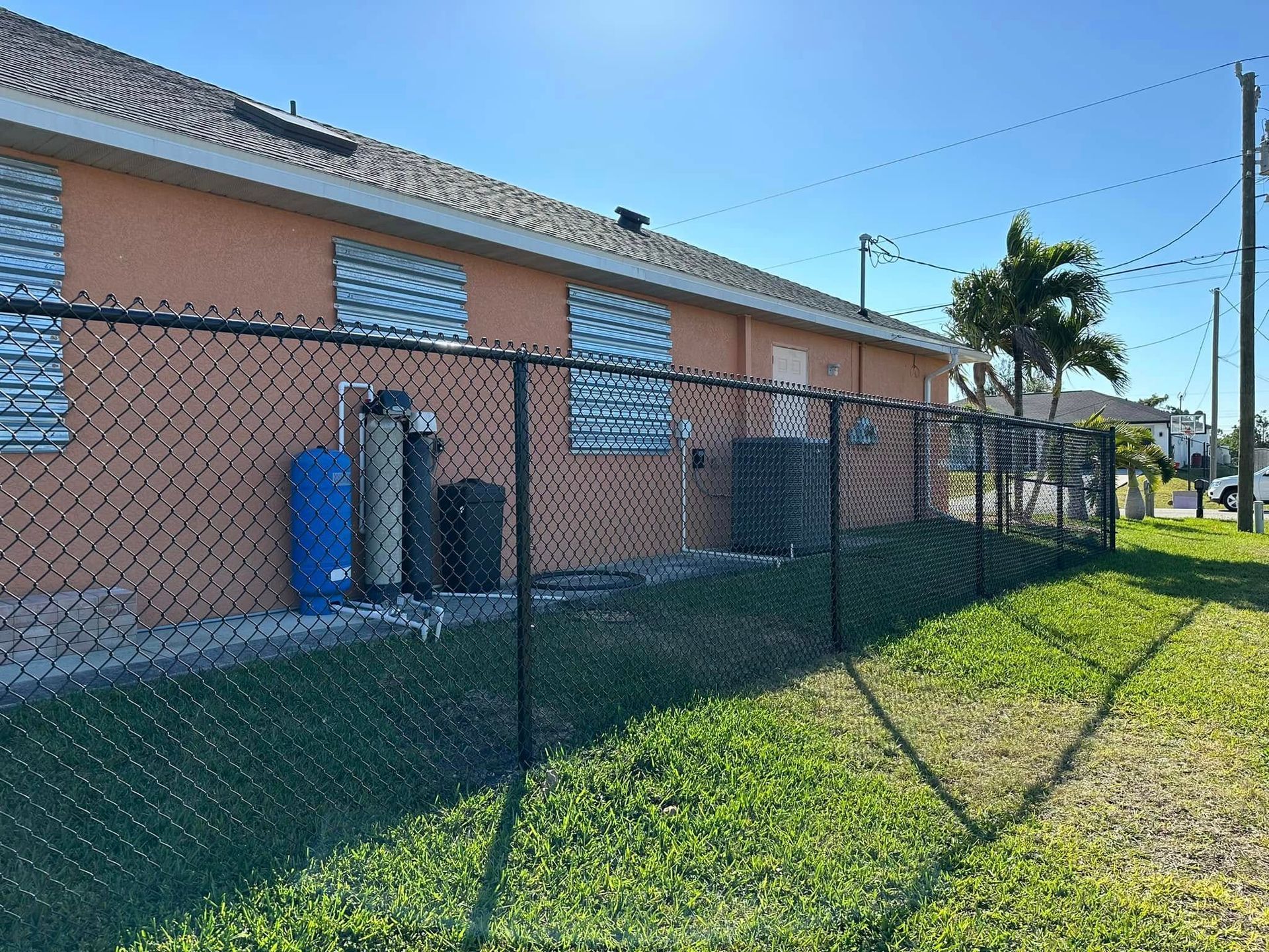 A building with boarded-up windows, behind a chain-link fence. Blue sky overhead, green grass in foreground.