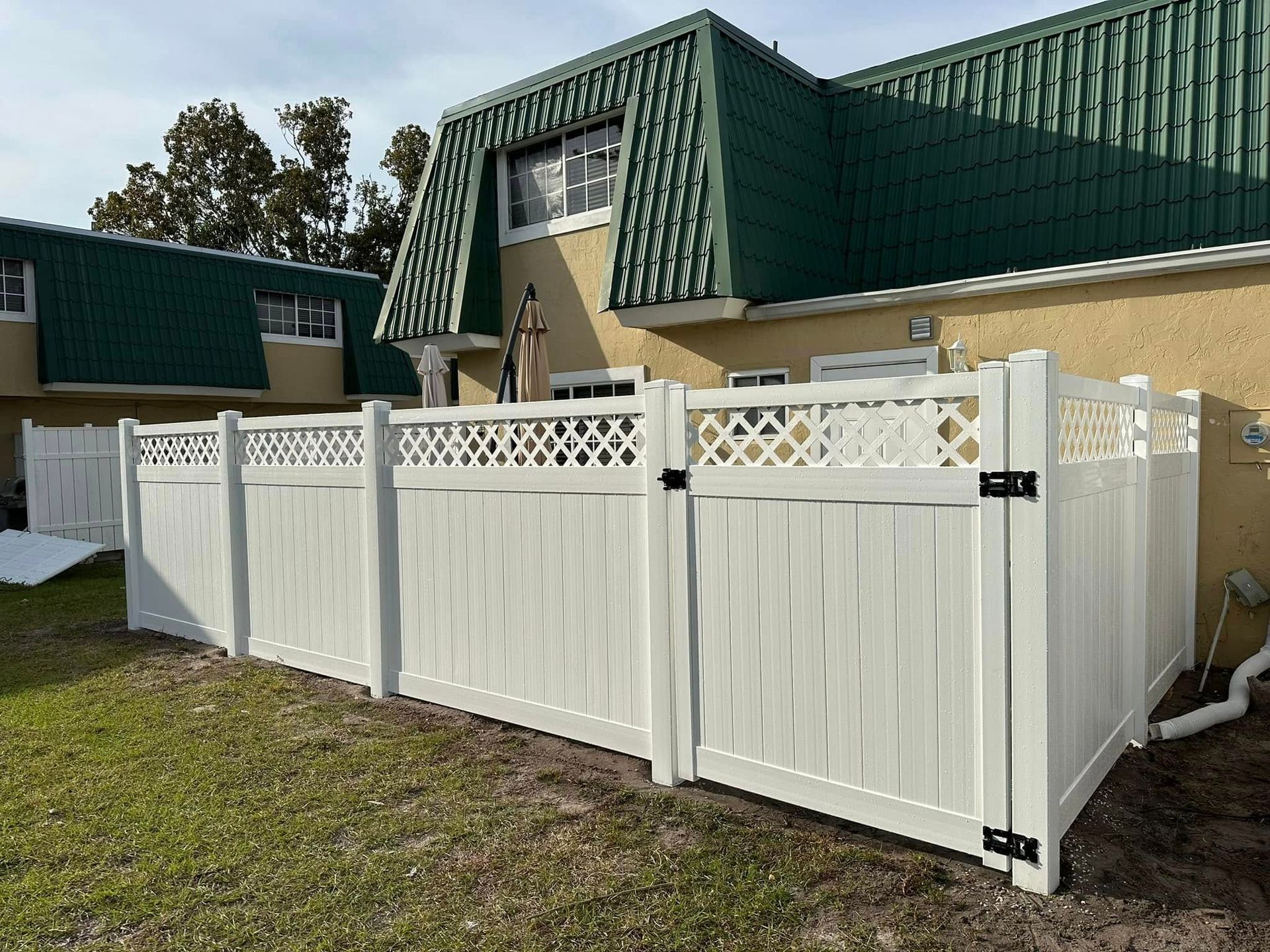 White vinyl fence encloses a yard, with a matching gate. Background shows a two-story building with green roof.