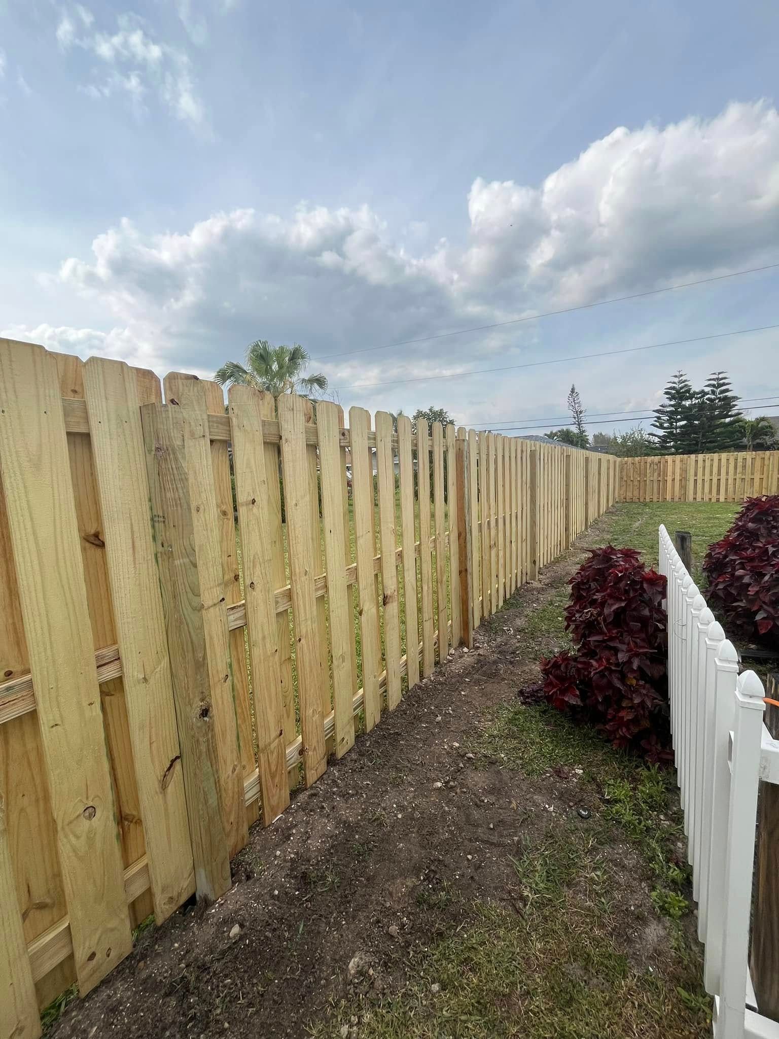 Wooden fence curves around a yard, bordering plants and grass under a cloudy sky.