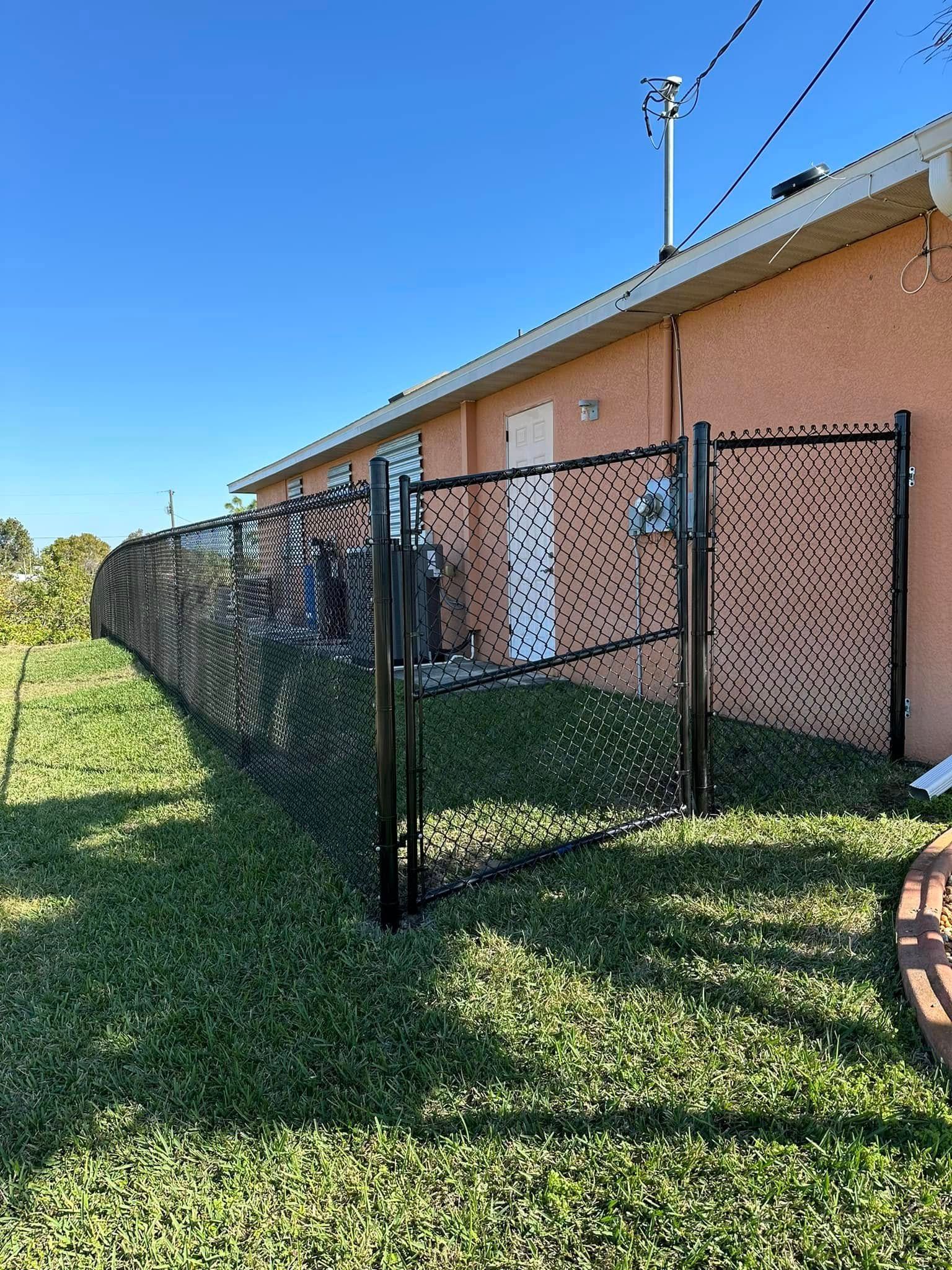 Black perforated metal fence with gate in grassy yard beside a light-orange building against blue sky.