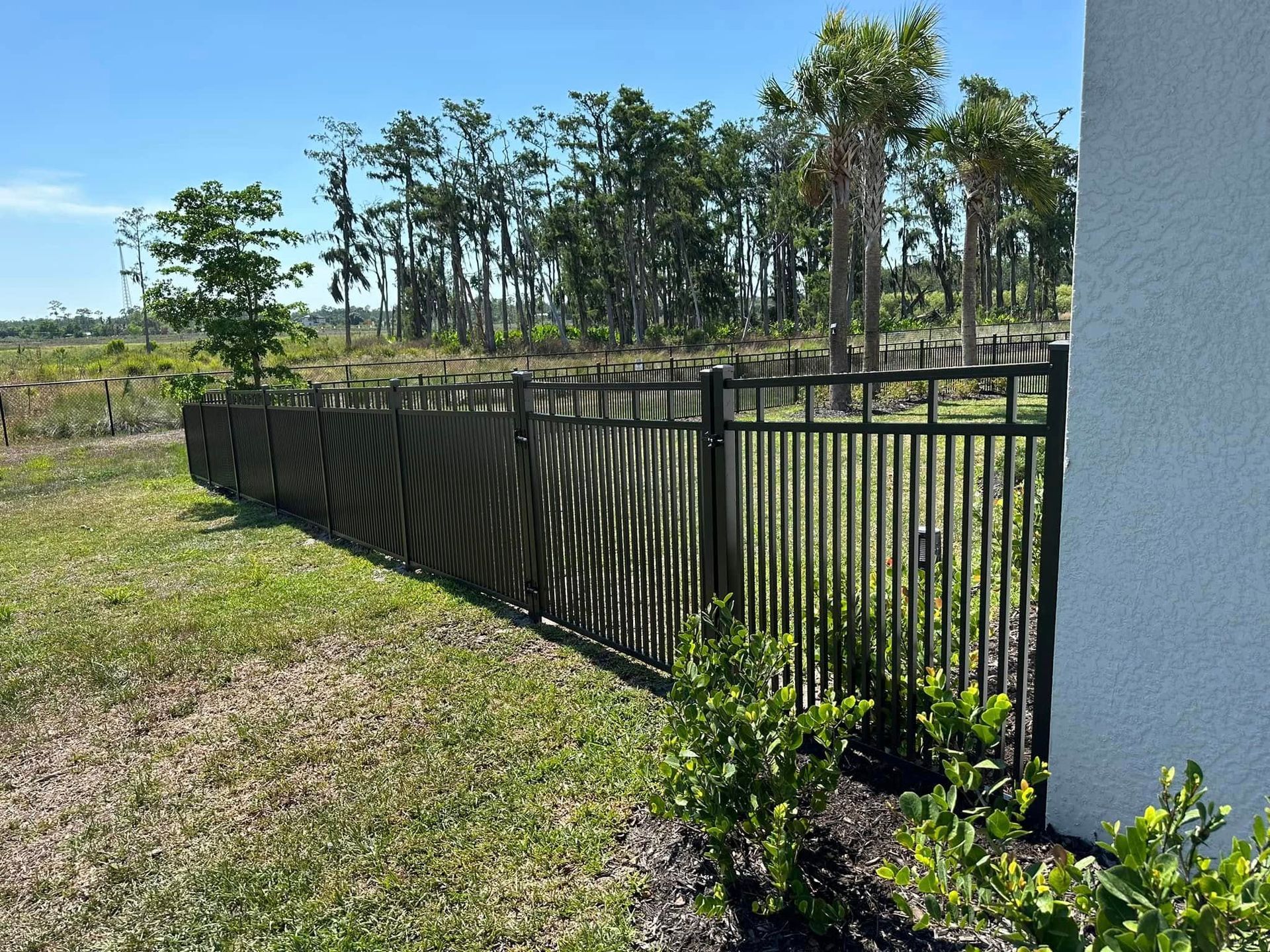 Black metal fence along grass, partial privacy screen, with trees in the distance and a white stucco wall.