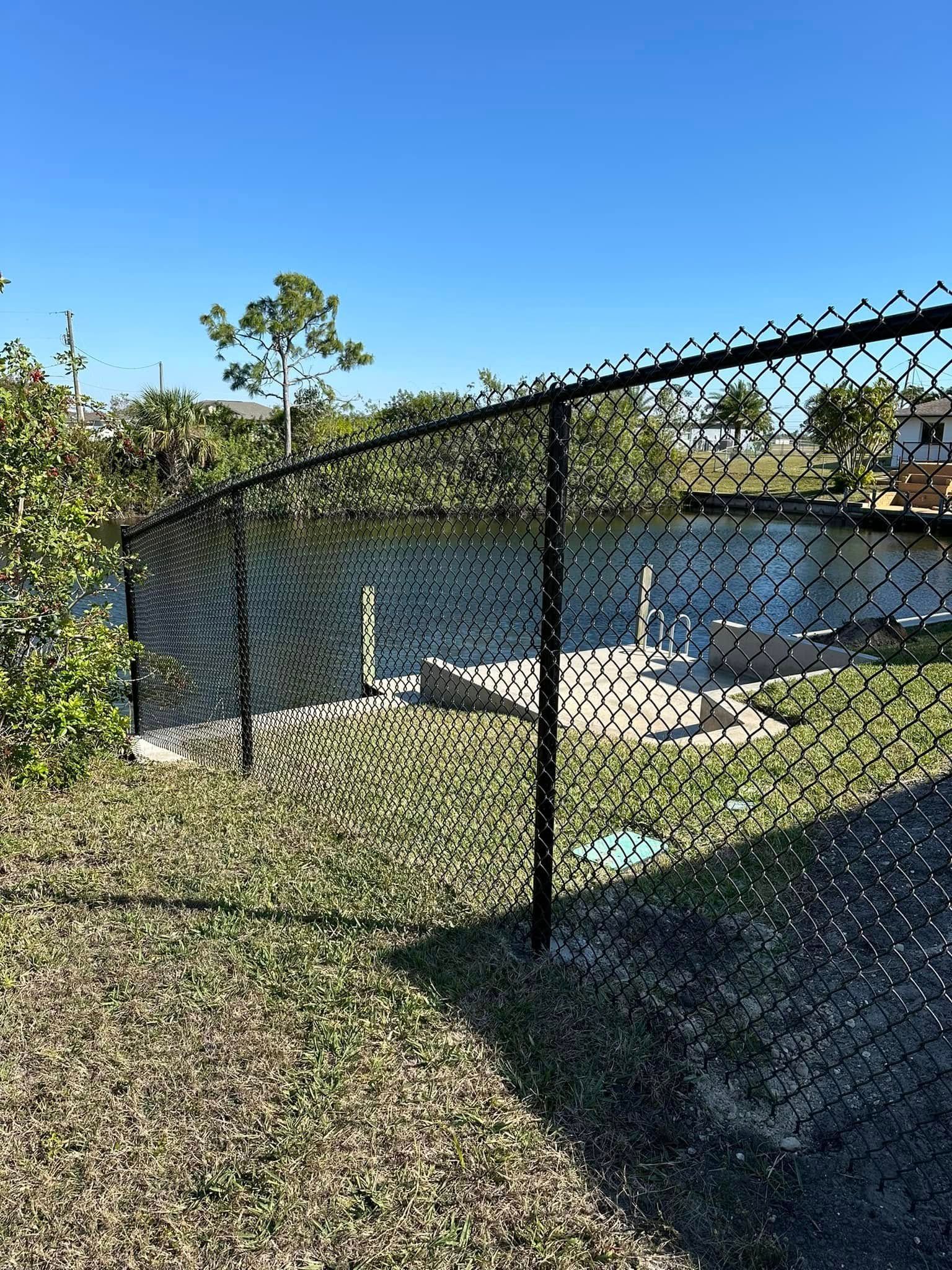 Black chain-link fence encloses a yard with a partially-visible structure and greenery under a blue sky.