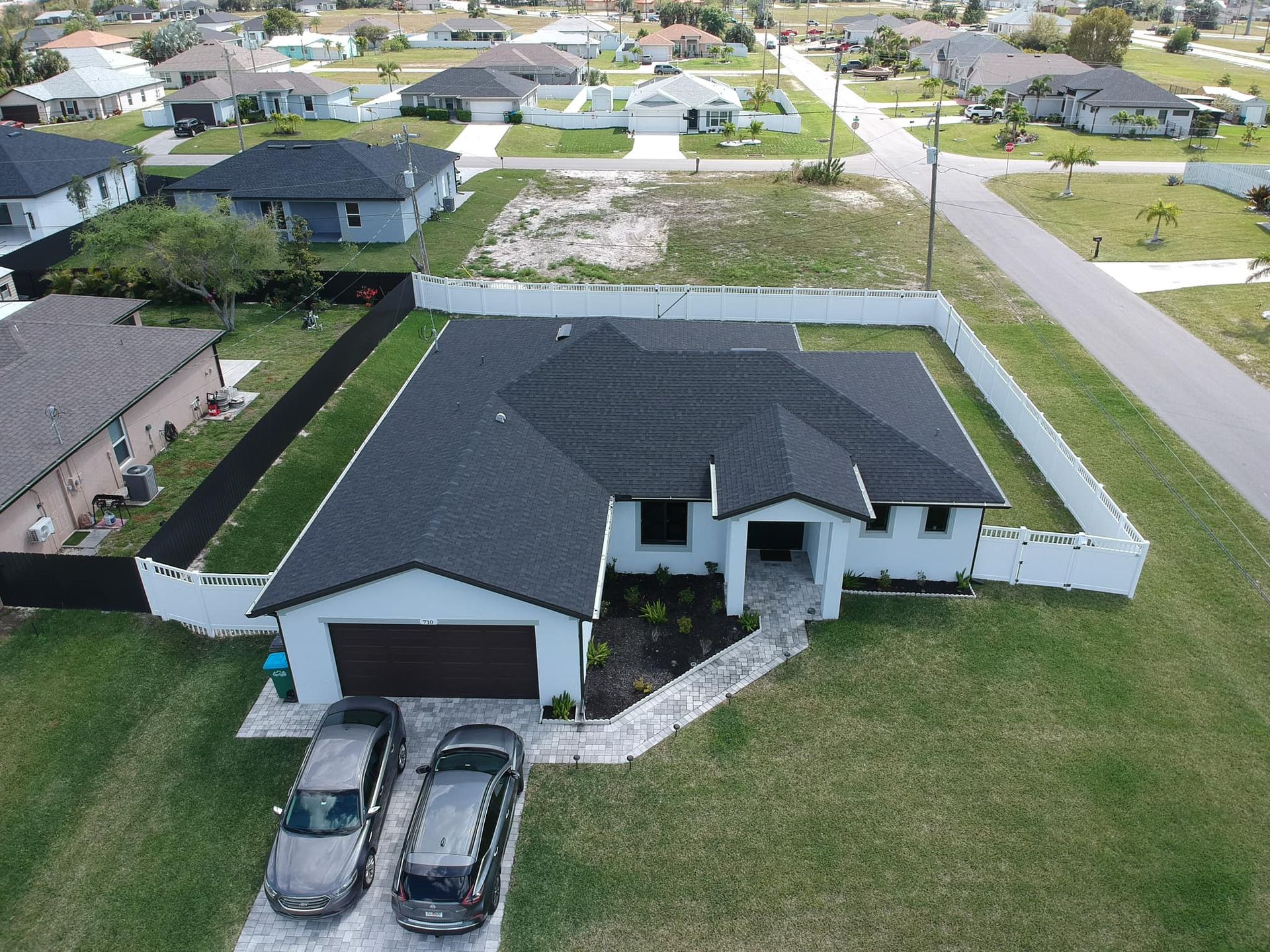 Aerial view of a white house with a dark roof, driveway with two cars, surrounded by a fence and grass.