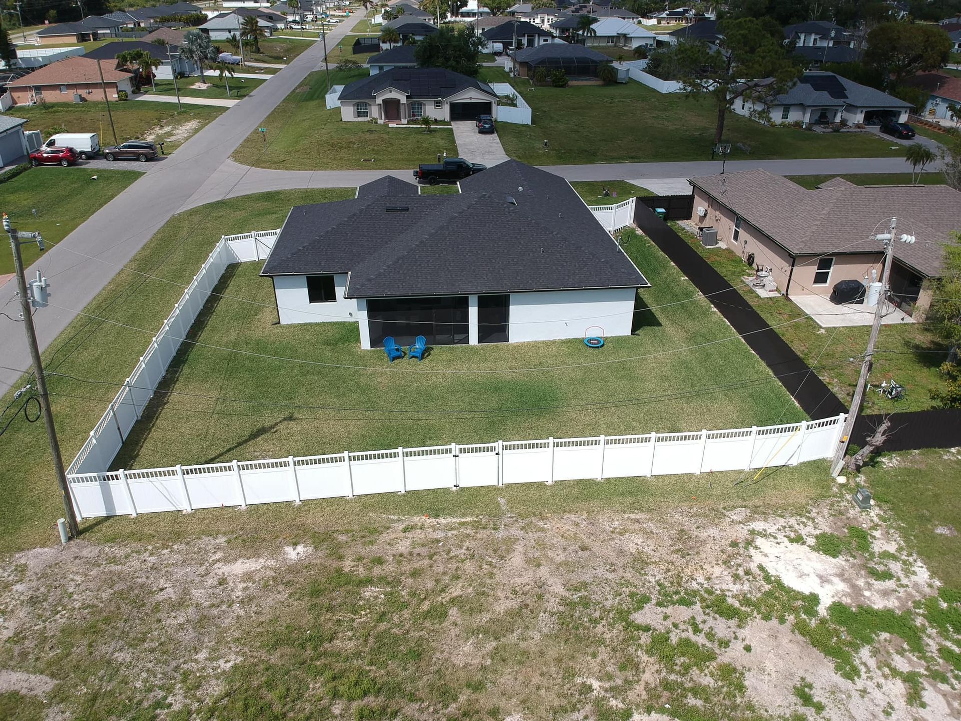 Aerial view of a house with a white fence, green lawn, and black roof in a residential area.