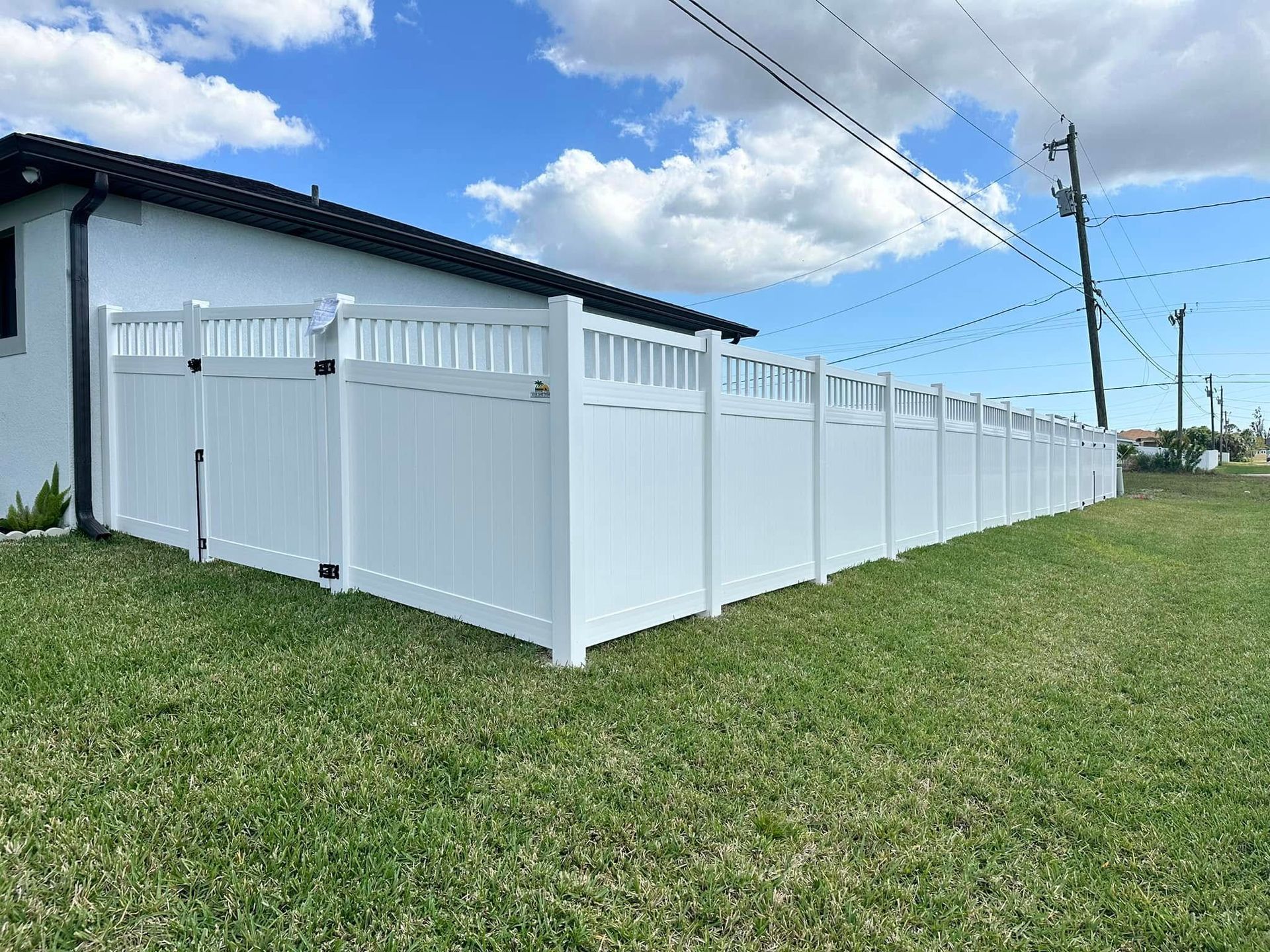 White vinyl fence around a house on a grassy lawn under a partly cloudy sky.