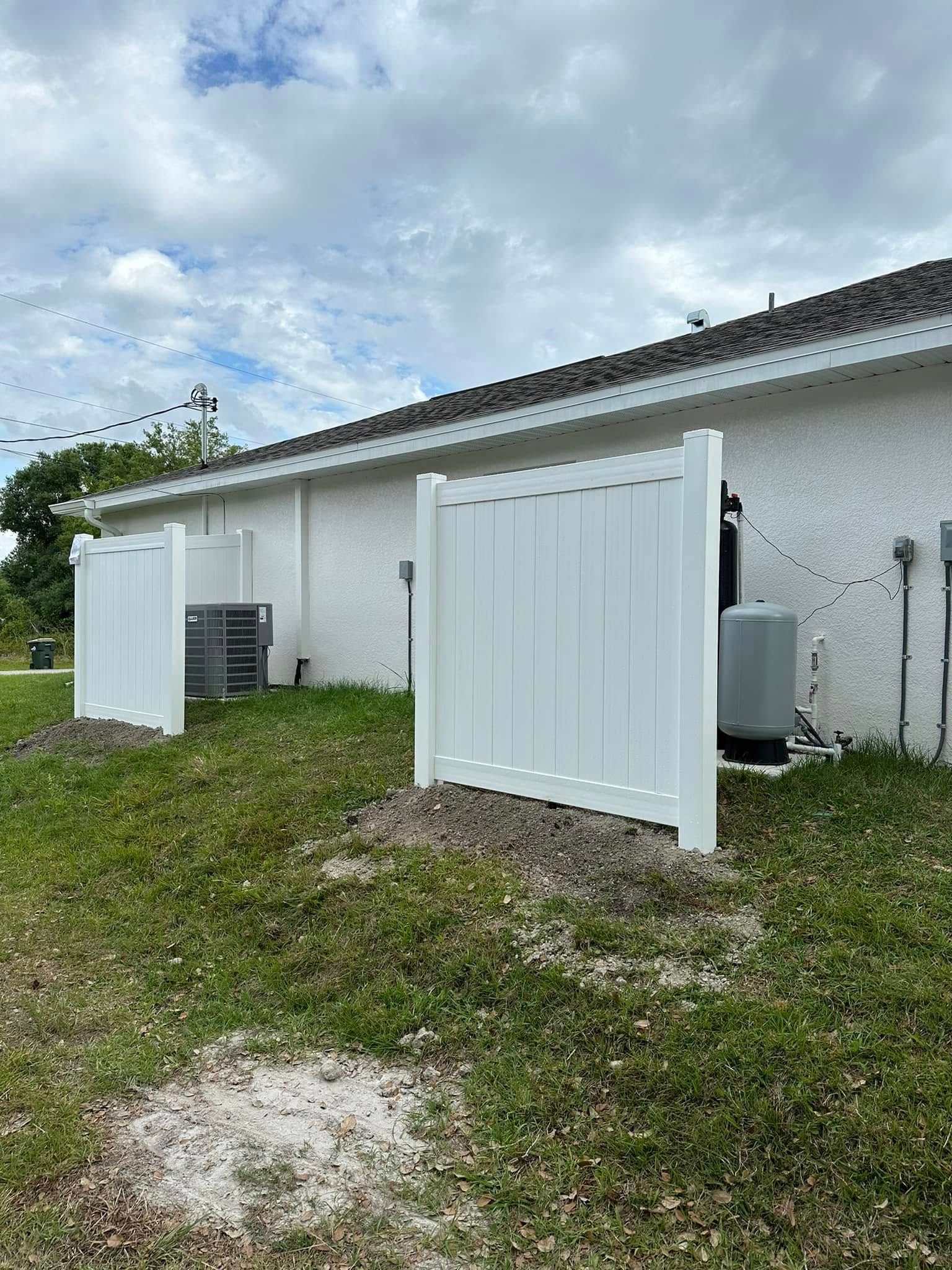 White privacy fences enclose HVAC equipment along the side of a light-colored house on a grassy lawn.