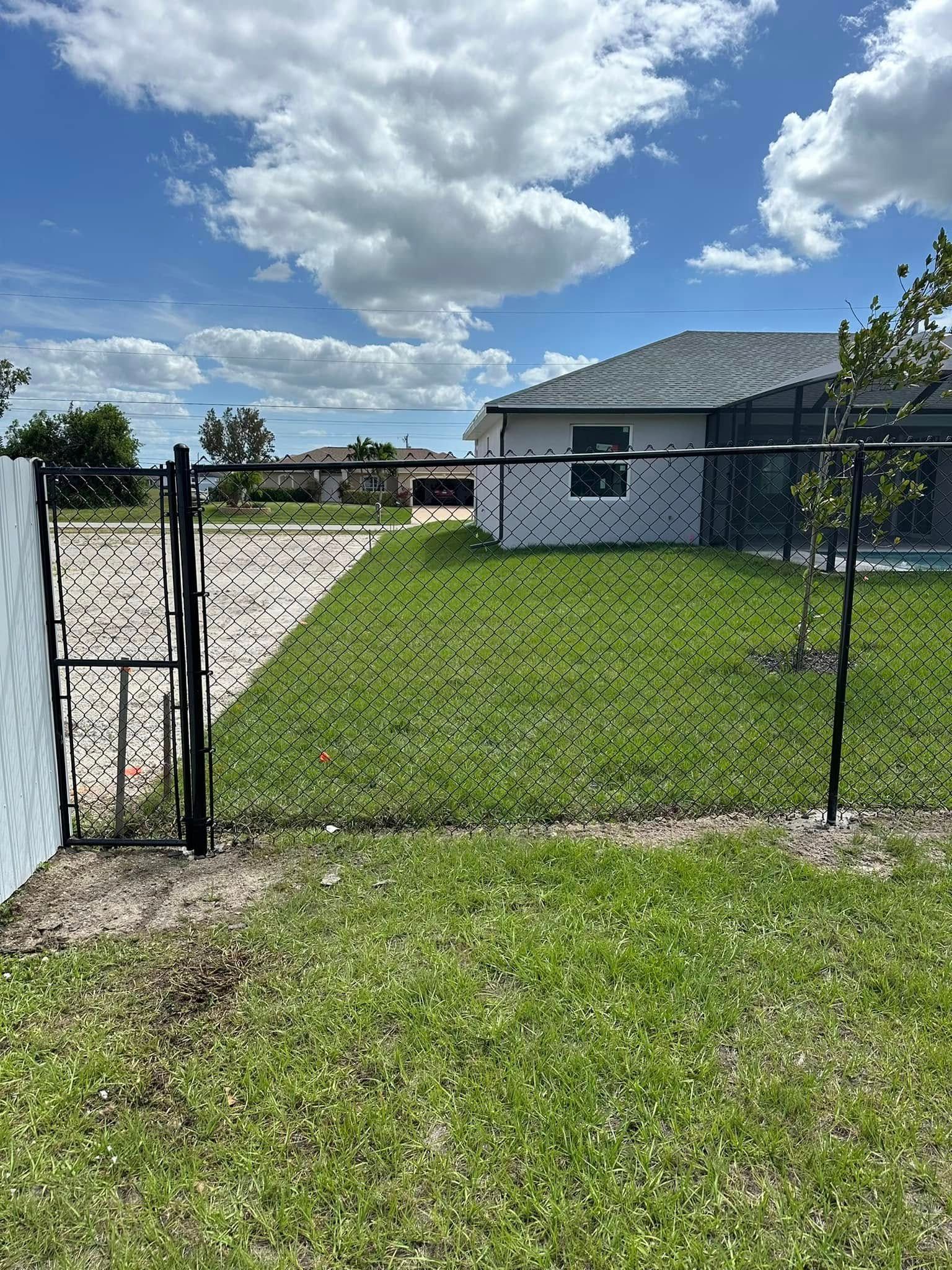 Black chain-link fence with gate, house in the background, green grass, blue sky with white clouds.