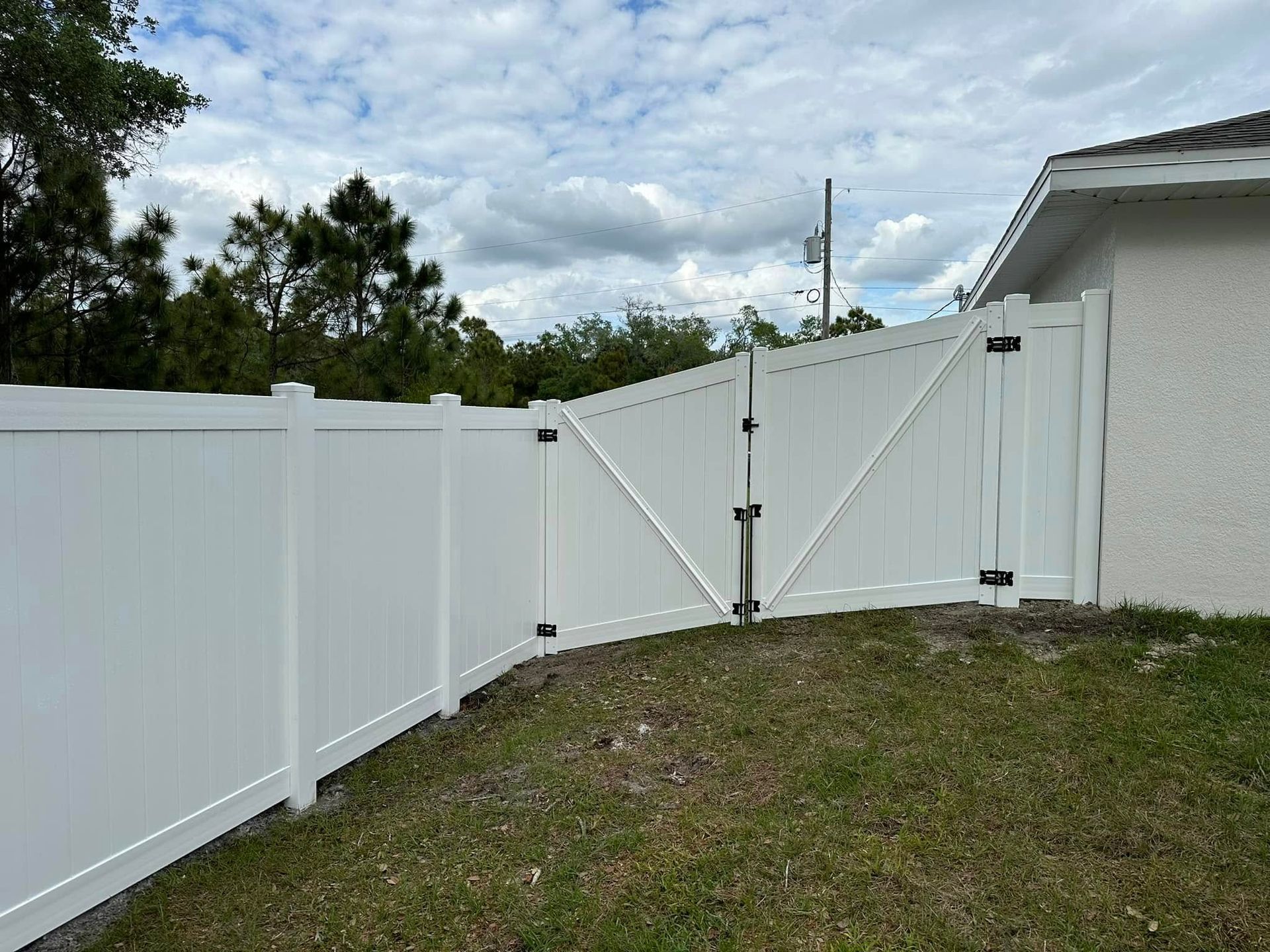 White vinyl fence with gate, set on grassy hill, against a cloudy sky and a house.