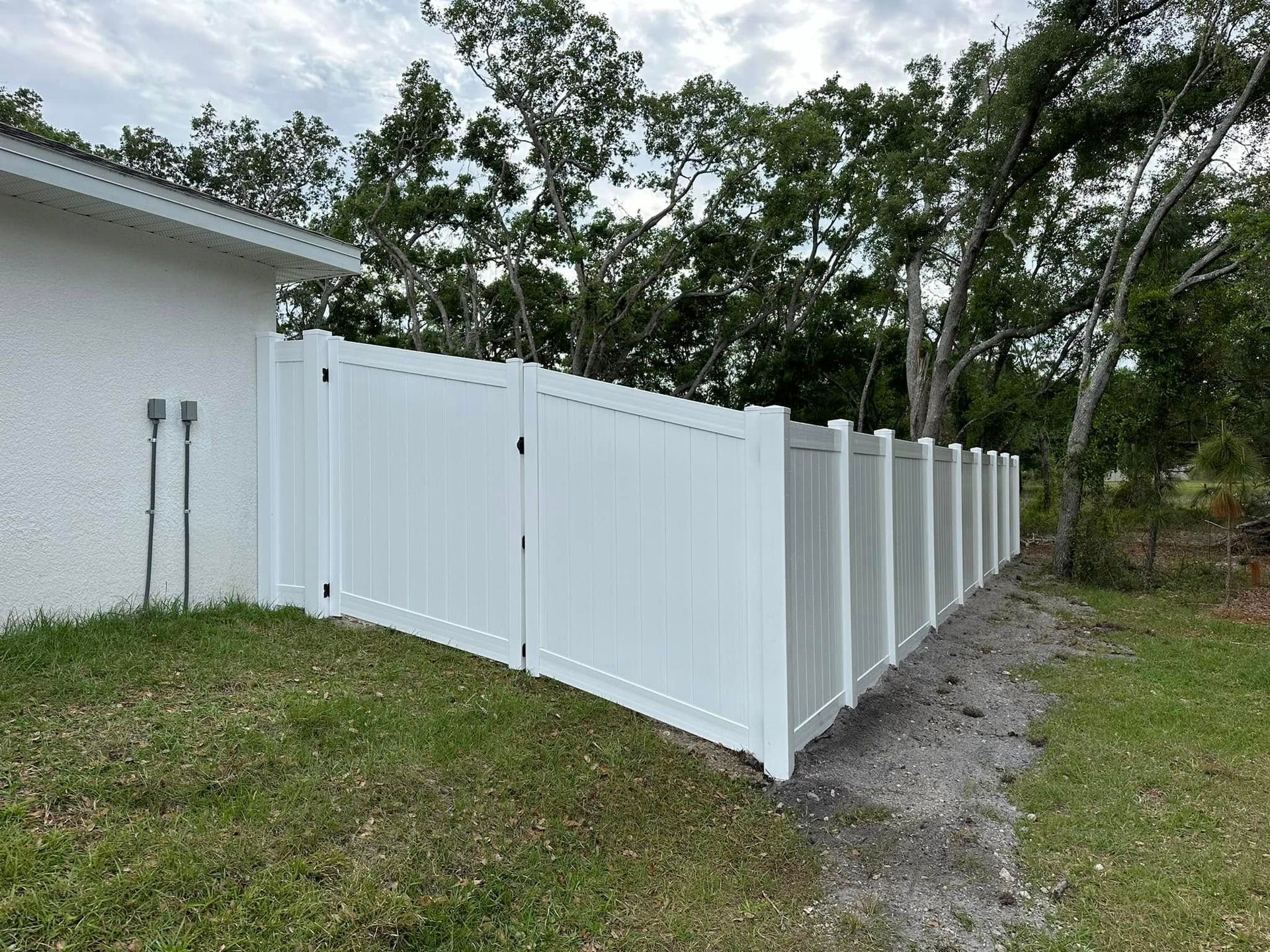 White vinyl fence bordering a grassy yard, alongside a house. Green trees in the background.