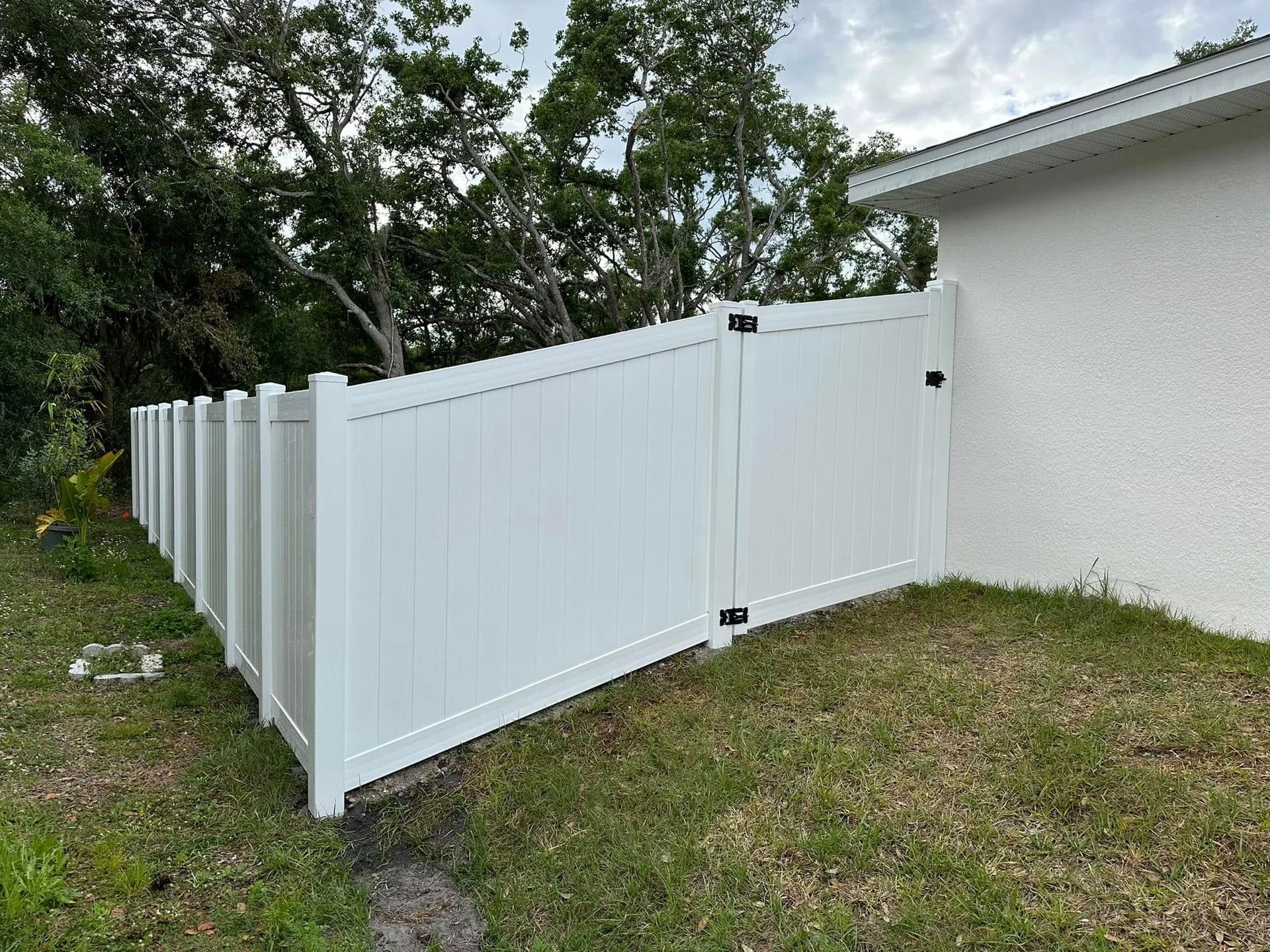 White vinyl fence with gate next to a house, green grass, and trees.