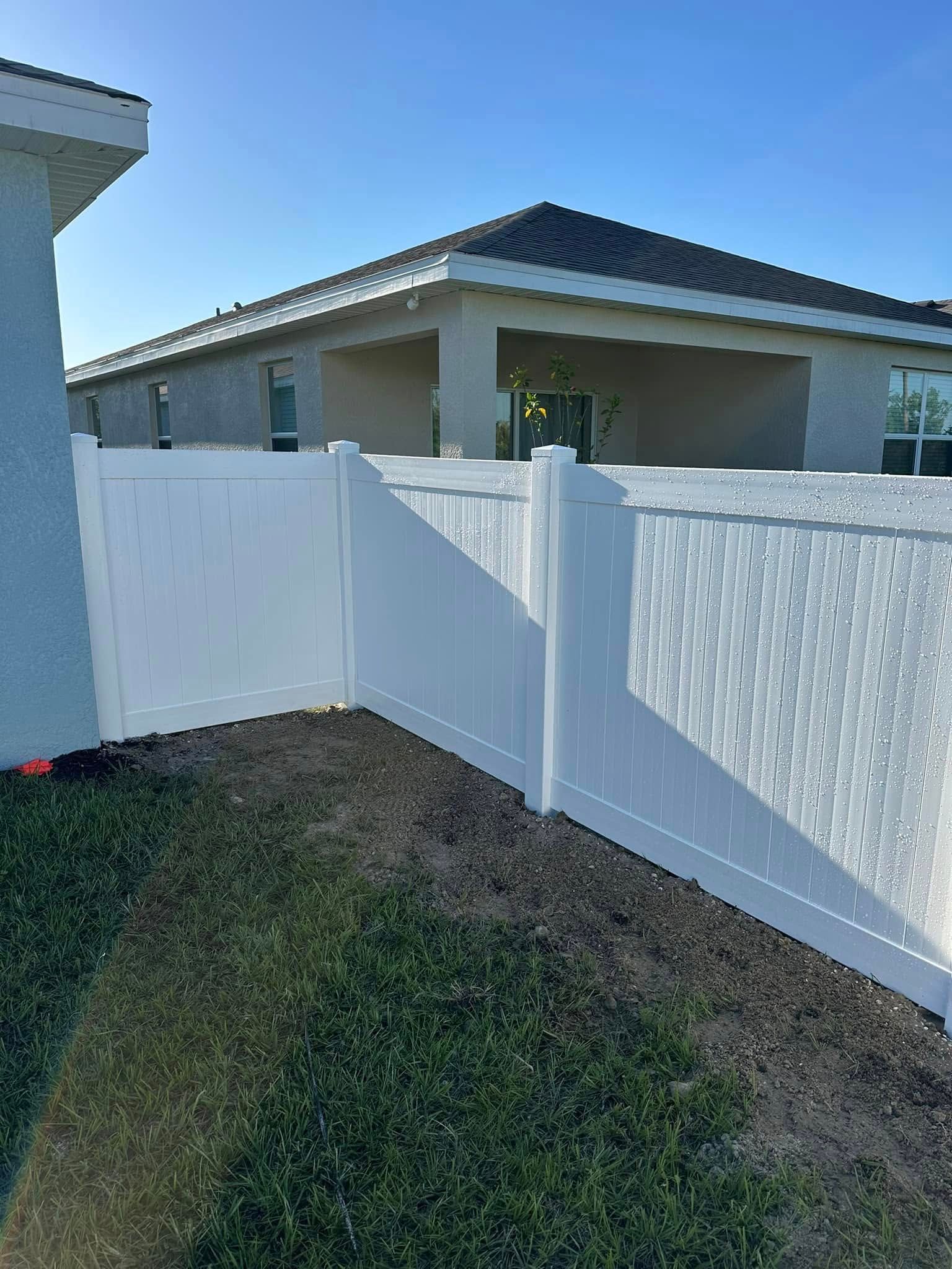 White vinyl fence enclosing a grassy yard, with a beige house in the background under a clear, blue sky.
