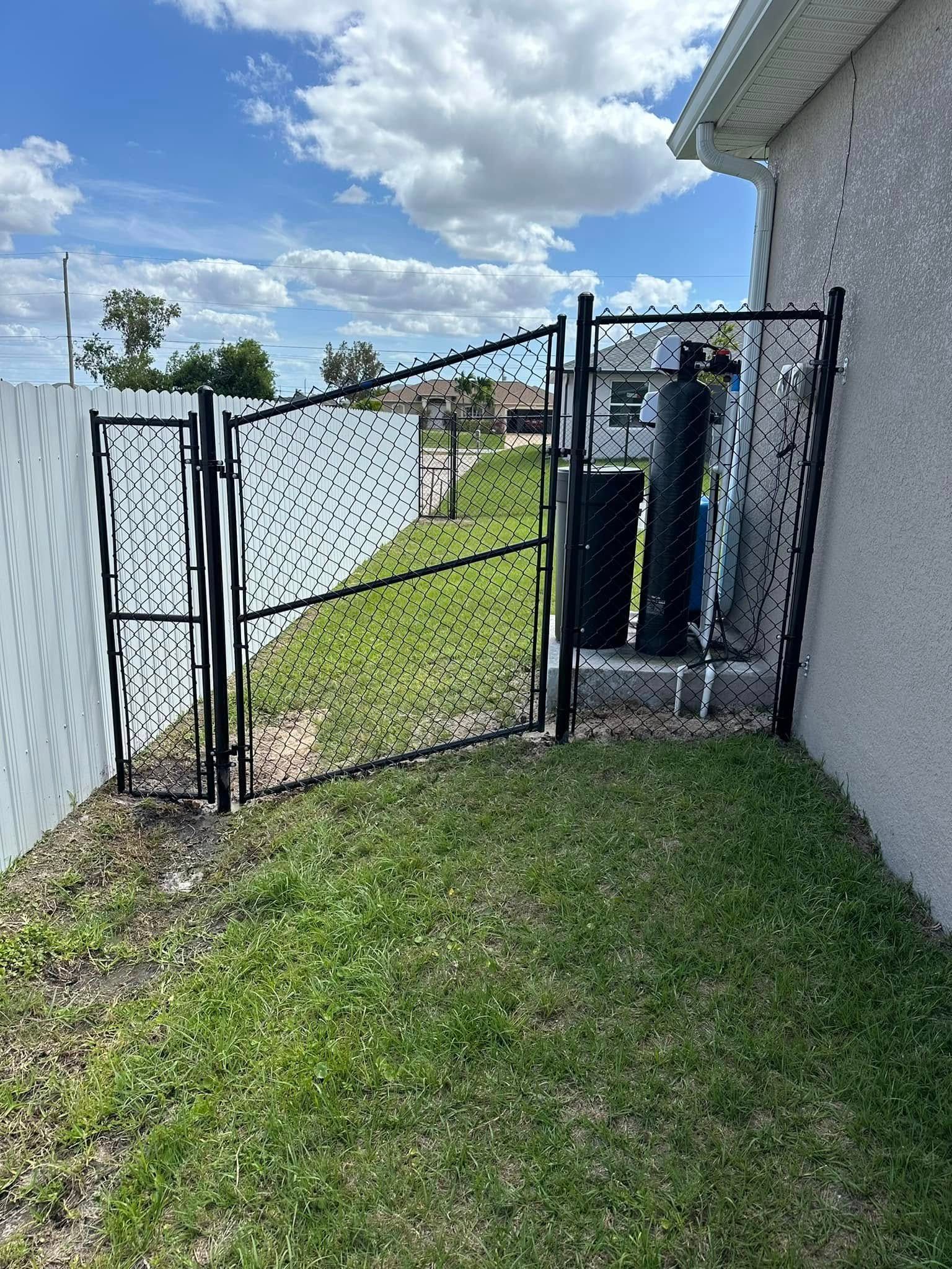 Black metal gate in a grassy area, leading to the side of a house with a cylindrical object next to it.