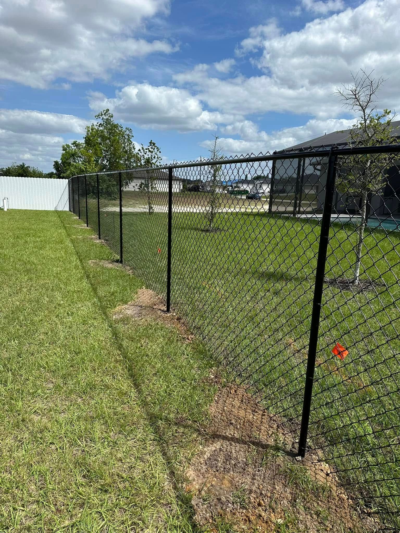 Black chain-link fence in grassy backyard against a blue sky with clouds.