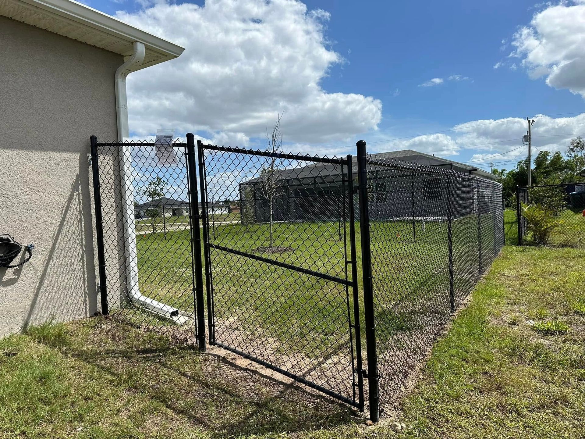 Black chain-link fence with gate, beside a beige house. Green grass and blue sky with clouds.