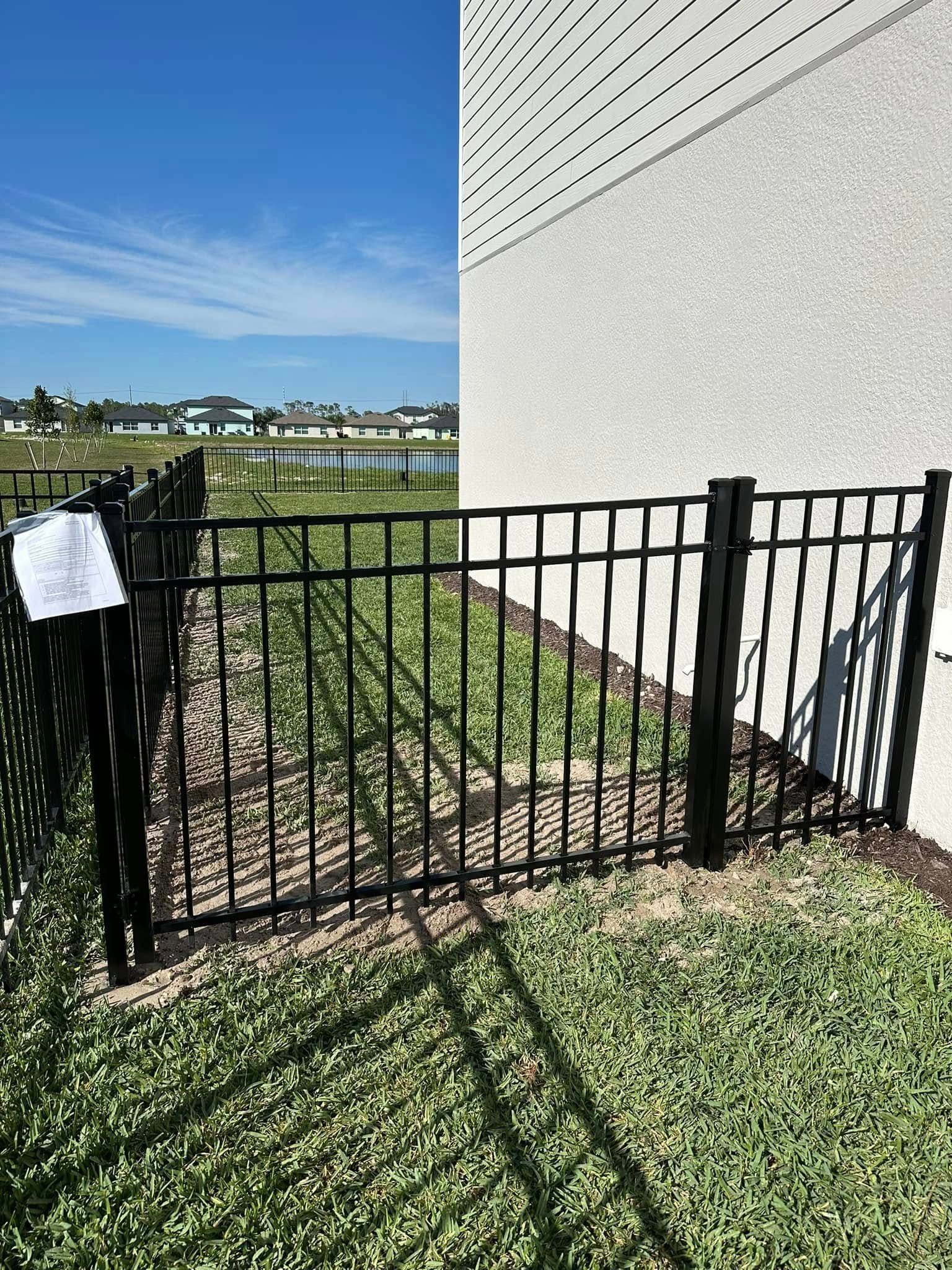 Black metal fence bordering a yard, next to a light-colored building and green grass, with a lake in the background.