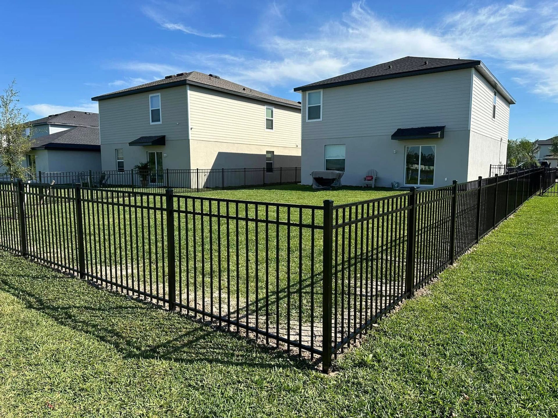 Black metal fence surrounds a grassy backyard with two-story houses under a blue sky.