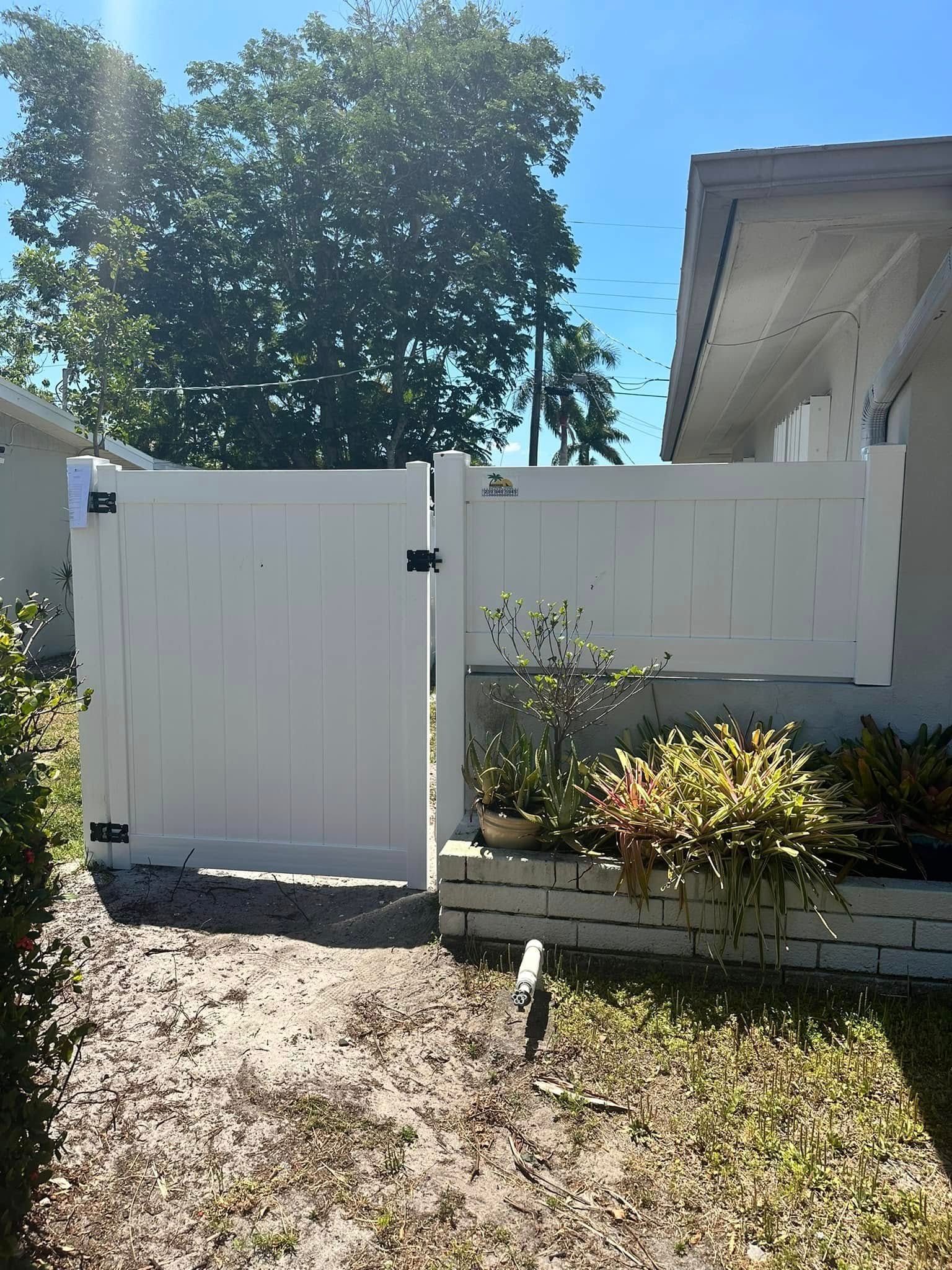 White vinyl fence with a gate in front of a house, with plants and a tree in the background.