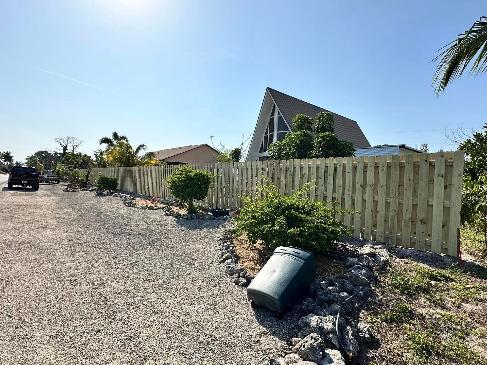 Wooden fence along a gravel driveway, leading to a building with a unique triangular roof, under a clear blue sky.