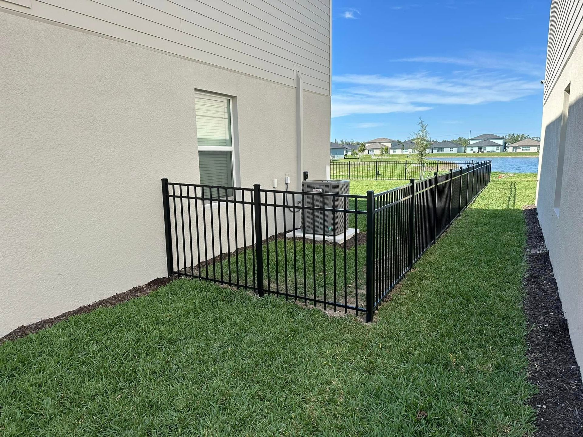 Black metal fence surrounds an AC unit between two light-colored buildings on a grassy lawn, with a lake in the background.