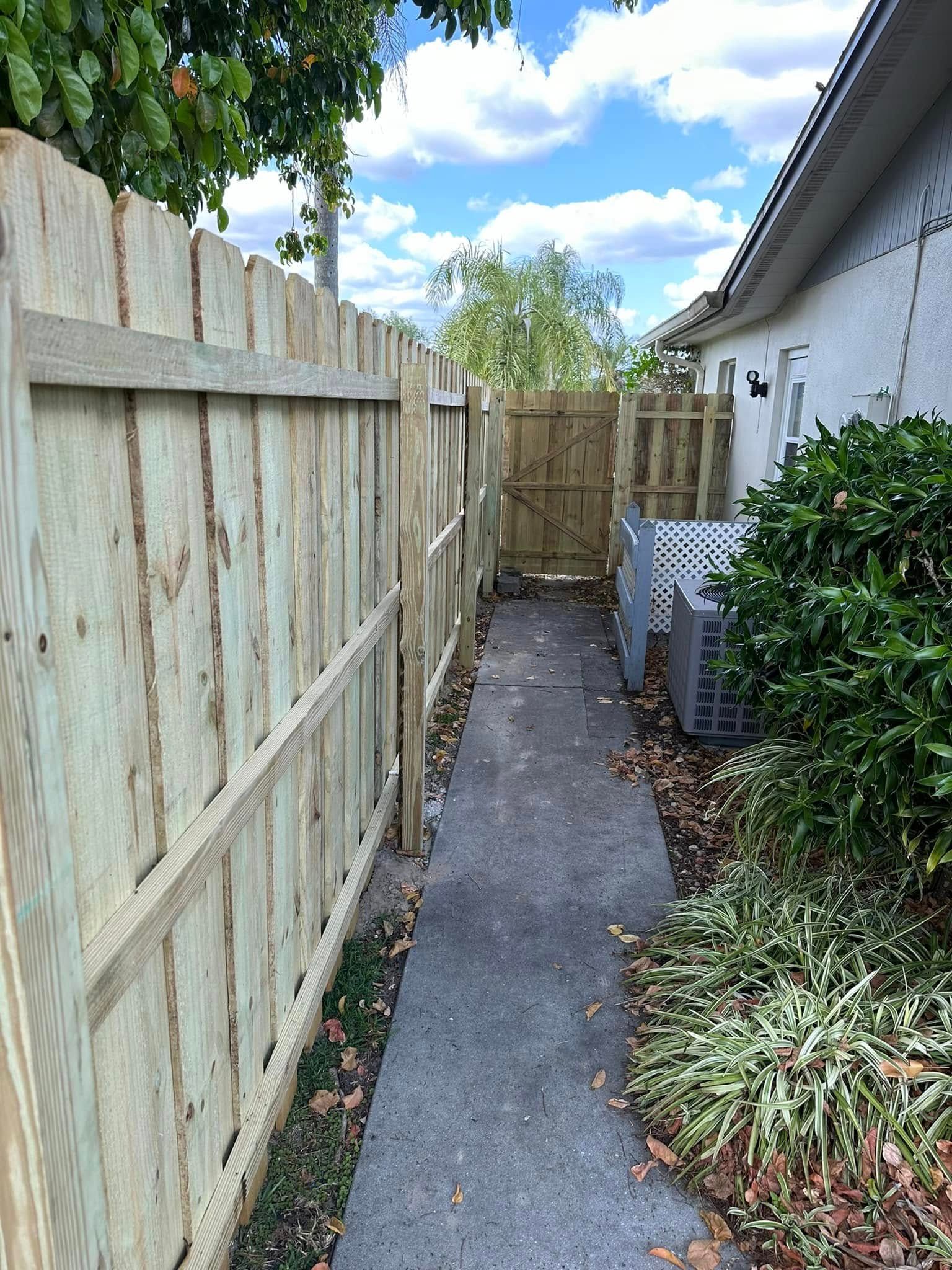 A narrow concrete walkway between a wooden fence and a building, with a gate at the end under a blue sky.