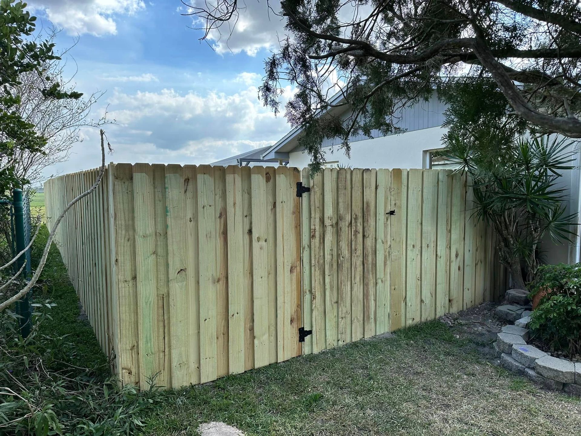 Wooden fence with a gate in a grassy yard.  Green grass and bushes, blue sky with clouds.
