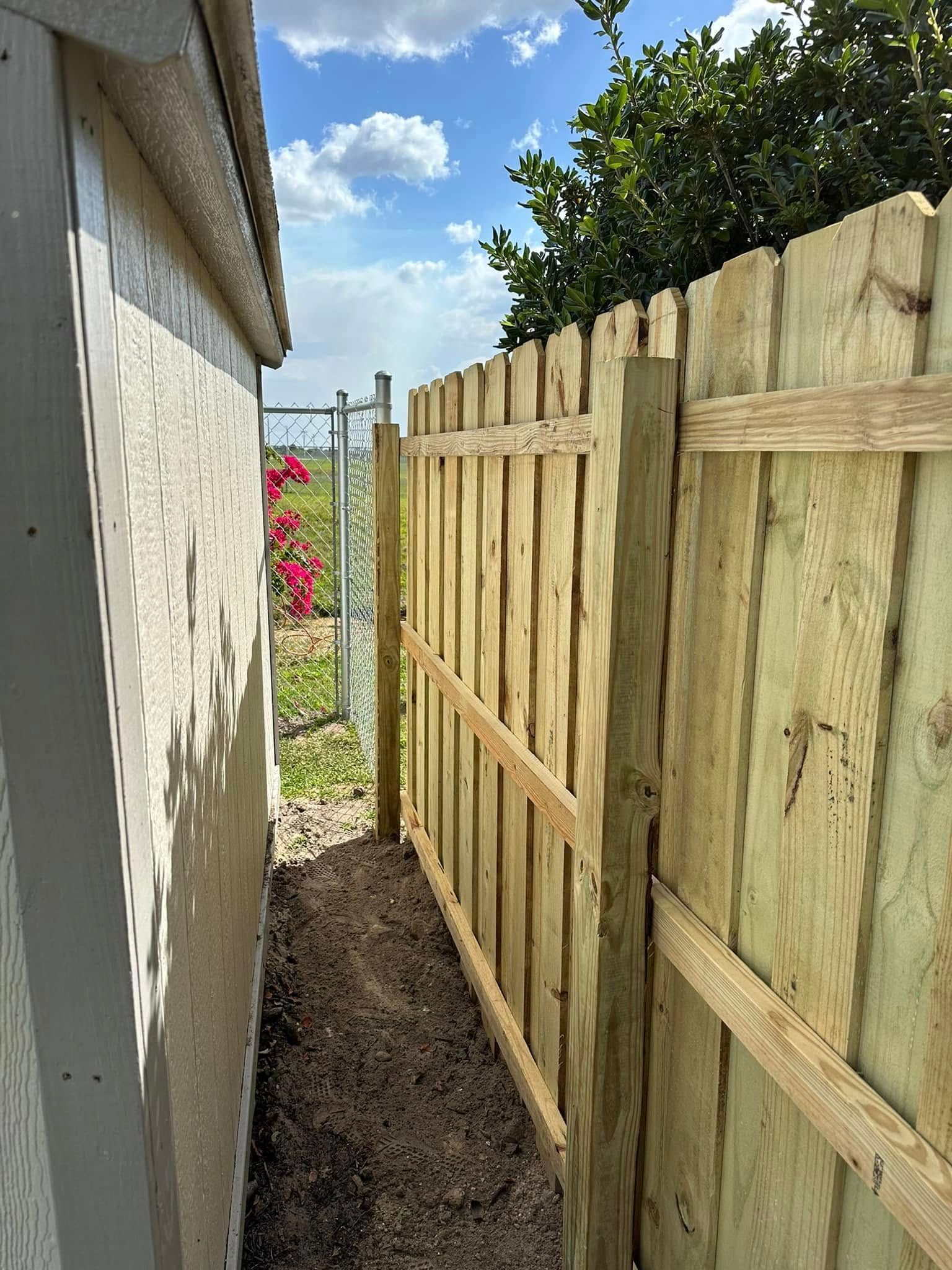 A wooden fence next to a building, with a dirt path in between. Bright sunlight.