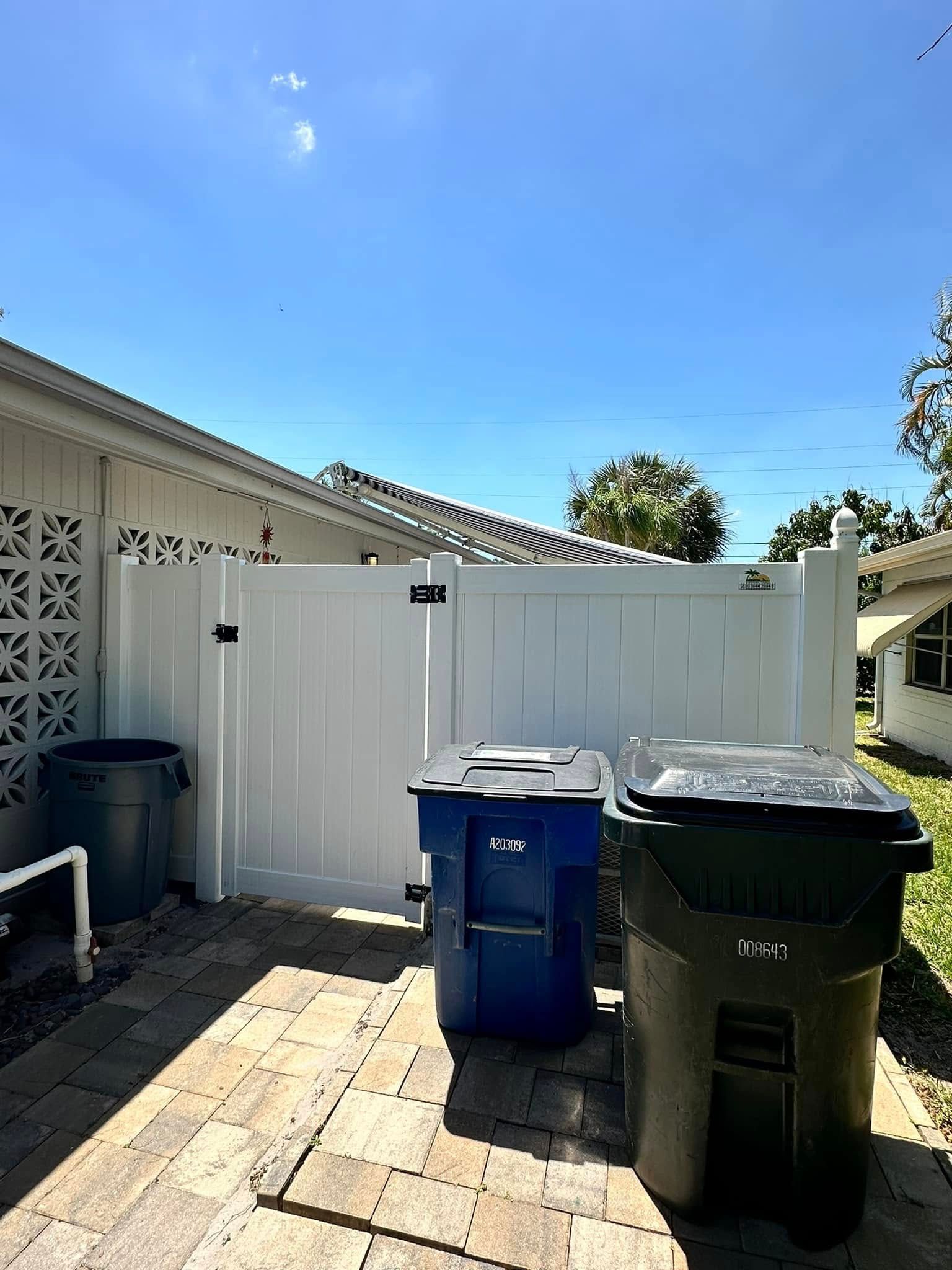 White fence with gate encloses three trash bins on a patio; sunny day.