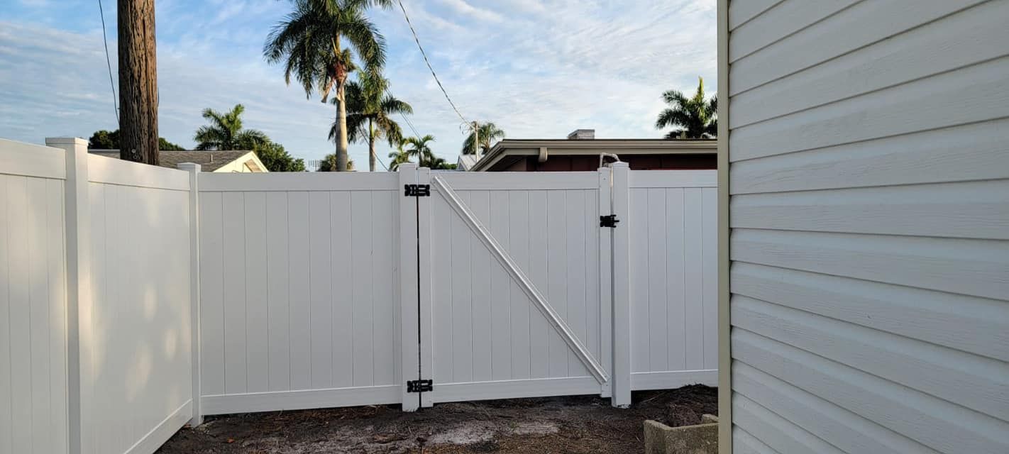 White vinyl fence and gate with a diagonal support, next to a white-sided building under a blue sky.