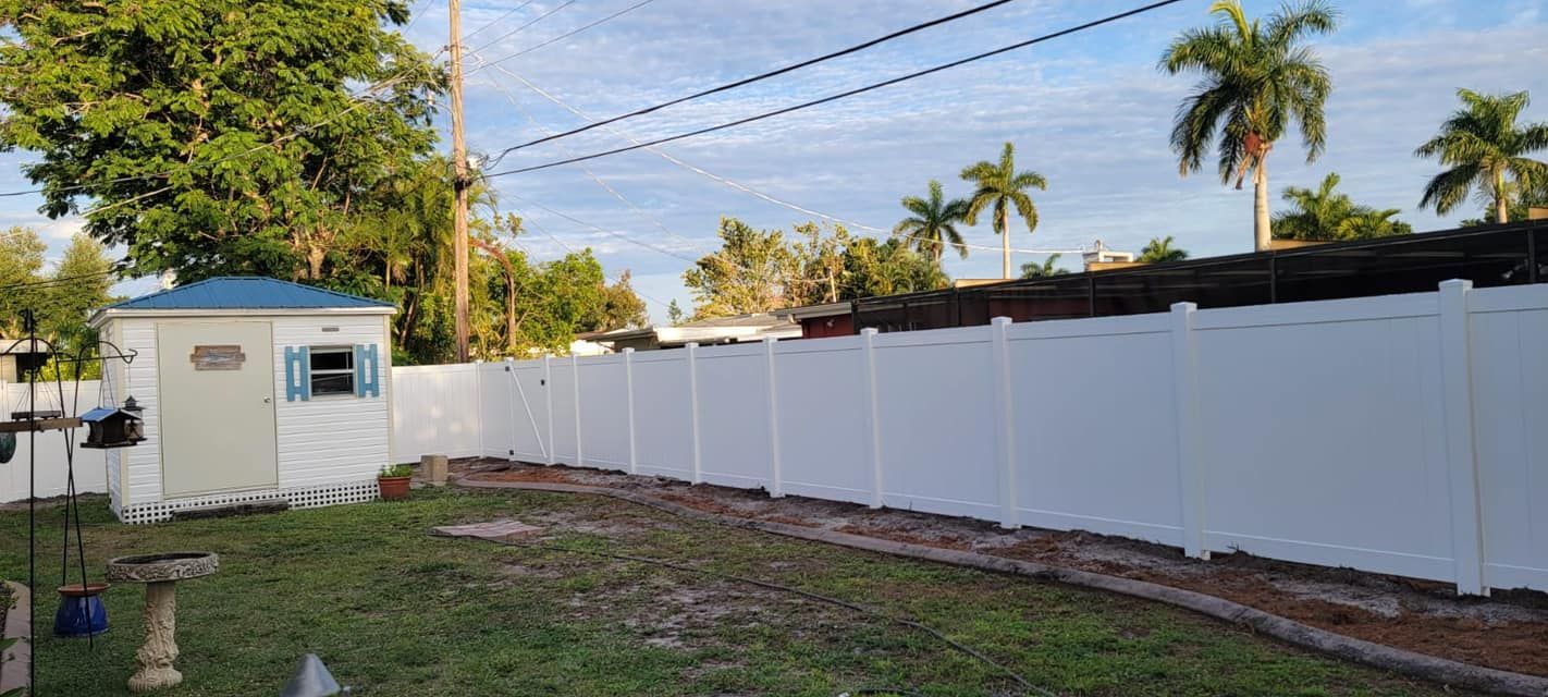 White privacy fence encloses a backyard with a shed, palm trees, and a partially cloudy sky.