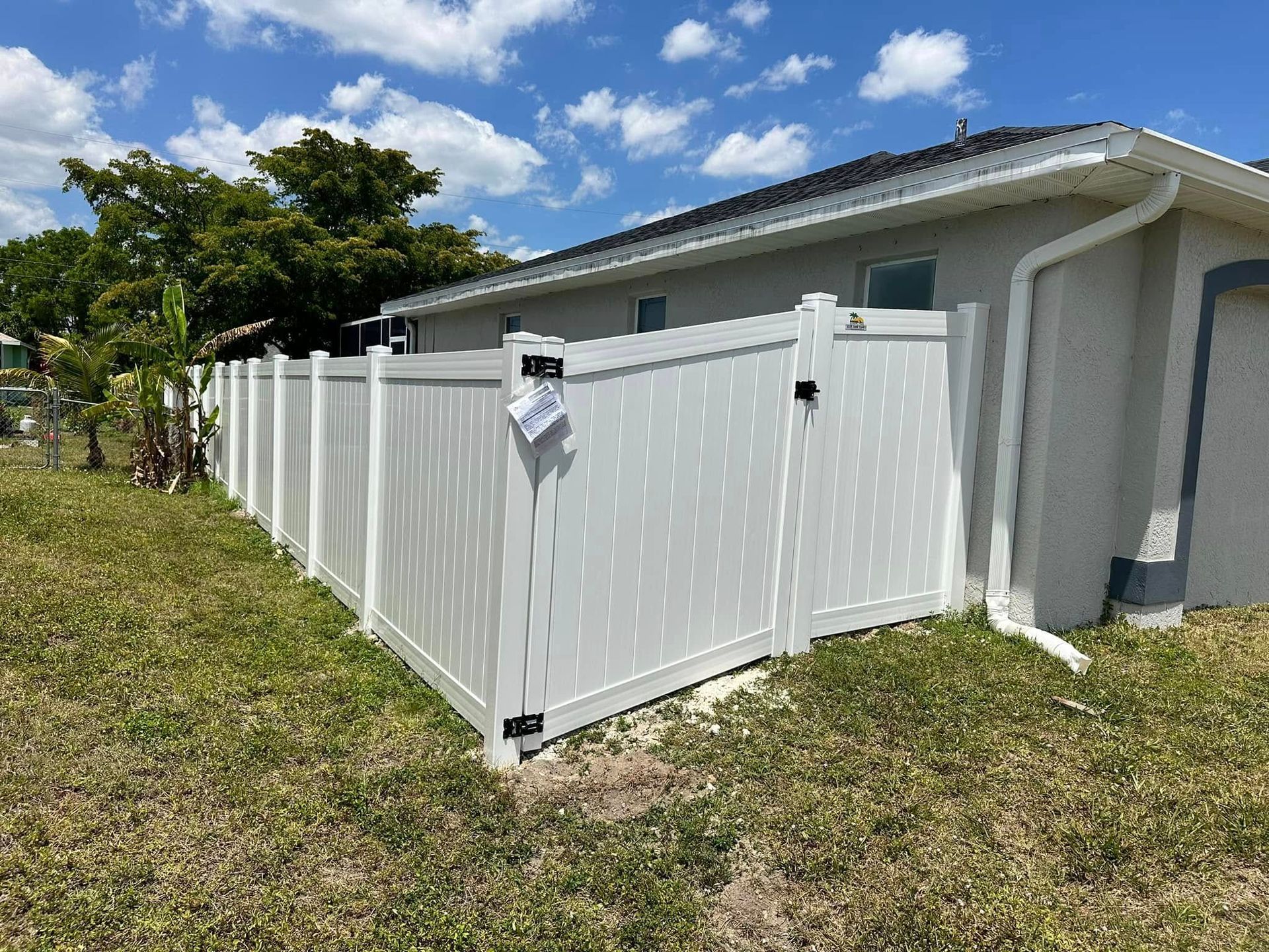 White vinyl fence with gate bordering a grassy yard next to a house under a cloudy blue sky.