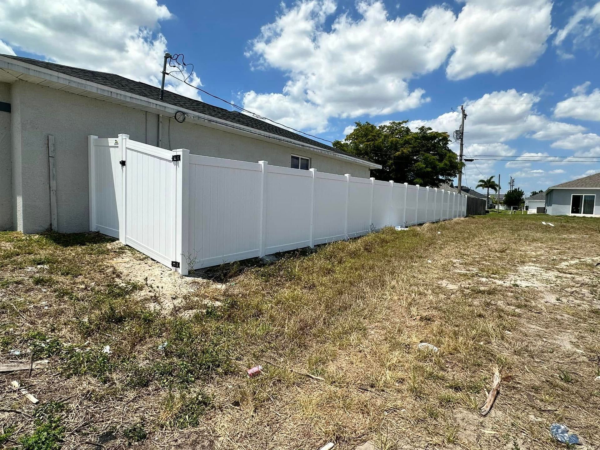 White vinyl fence along a house and yard, blue sky with clouds in background.